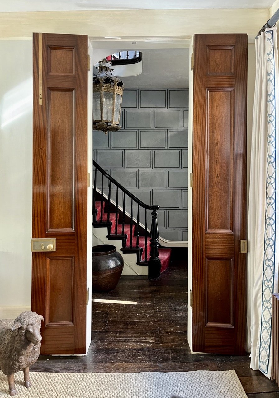 A view through wooden double doors into a hallway with a staircase, a large pot, and a hanging lantern.