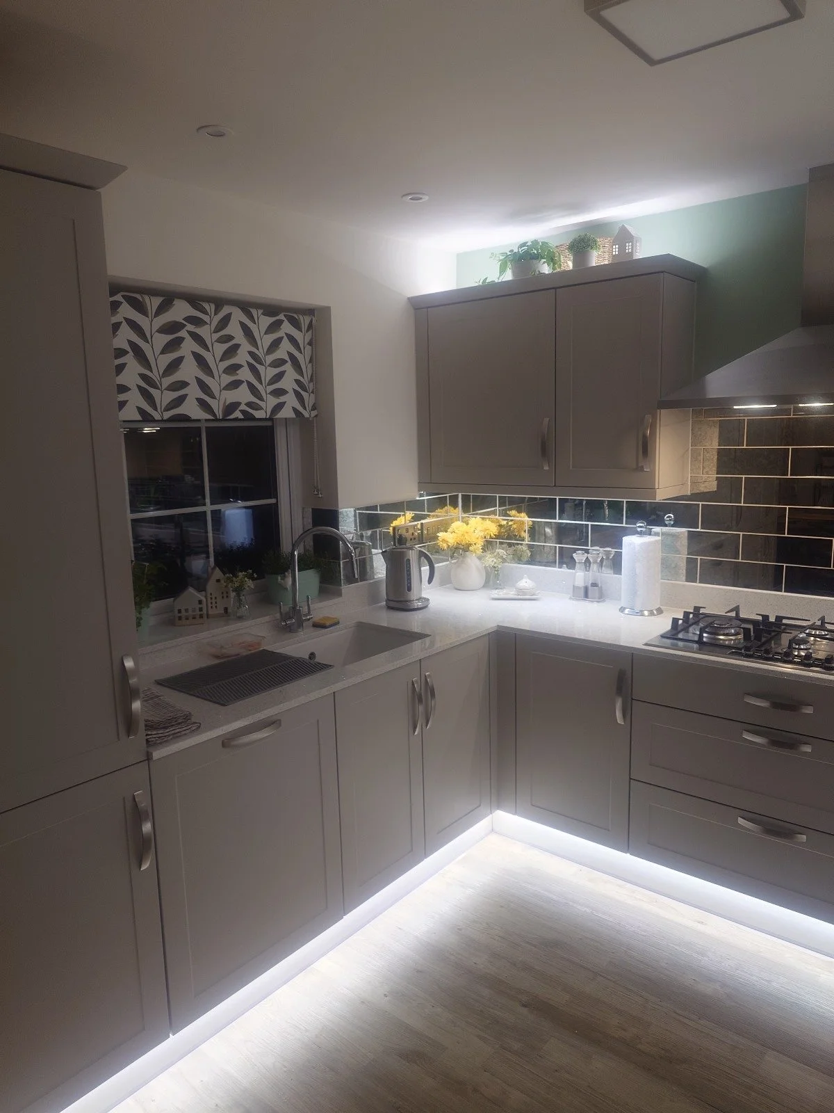 Modern kitchen with gray cabinets, stainless steel appliances, under-cabinet lighting, and decorative plants and flowers on the counter.