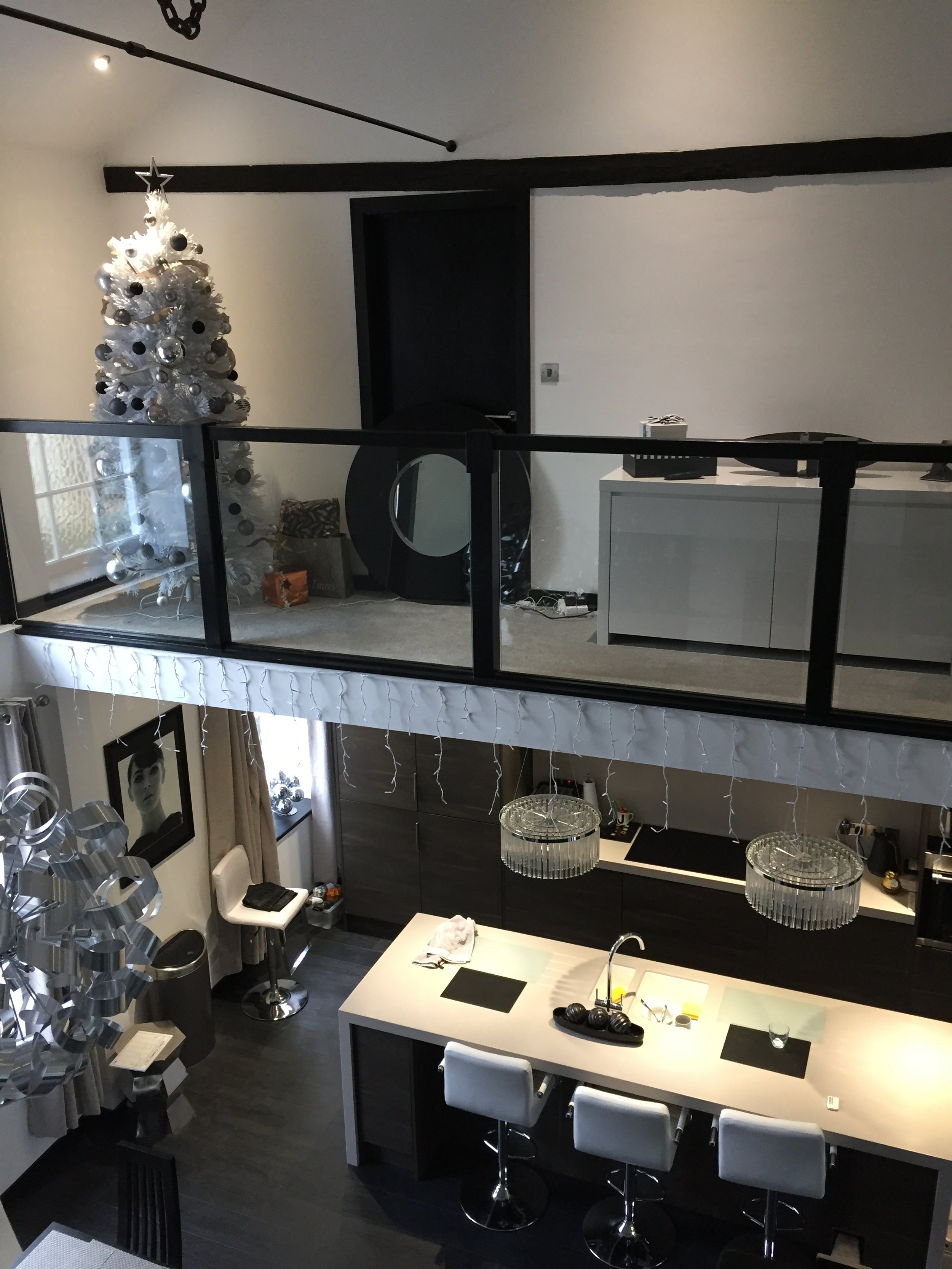 Modern open-plan kitchen and mezzanine with a decorated Christmas tree. The kitchen features a large island with white chairs, pendant lights, and a sleek design. A large metallic sculpture hangs from the ceiling. The mezzanine has a white desk and c