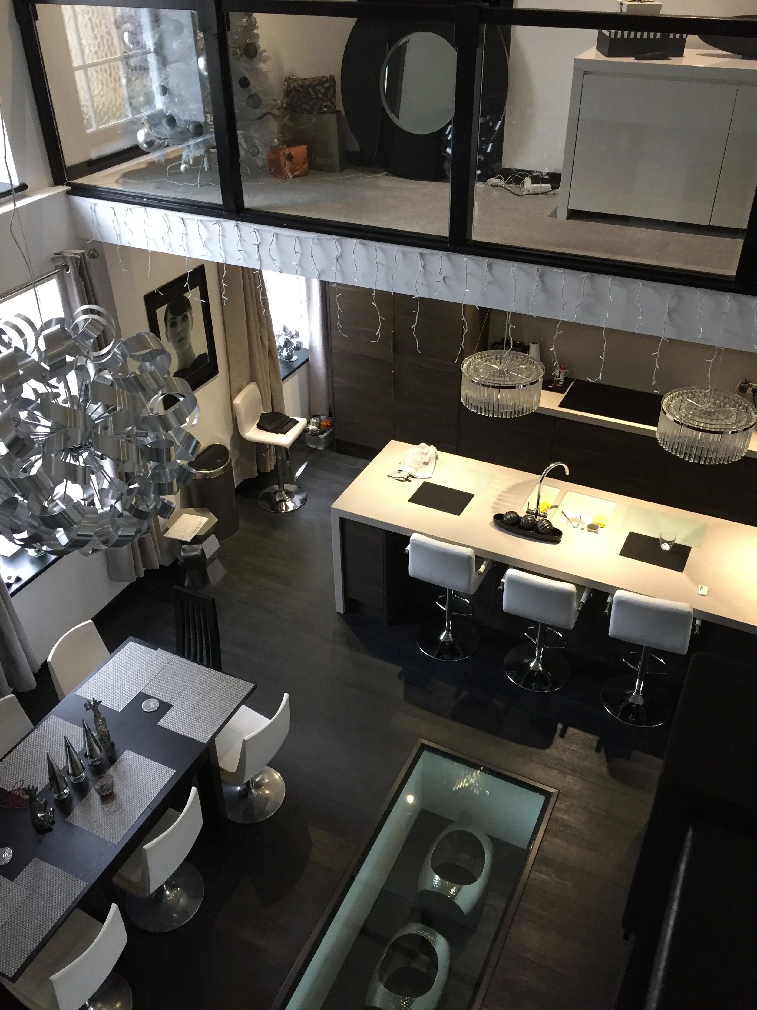 Modern loft-style living space with black and white decor, featuring a large kitchen island with bar stools, a dining table with white chairs, a unique chandelier, and a glass floor section. Upstairs, a small room with a white Christmas tree and deco