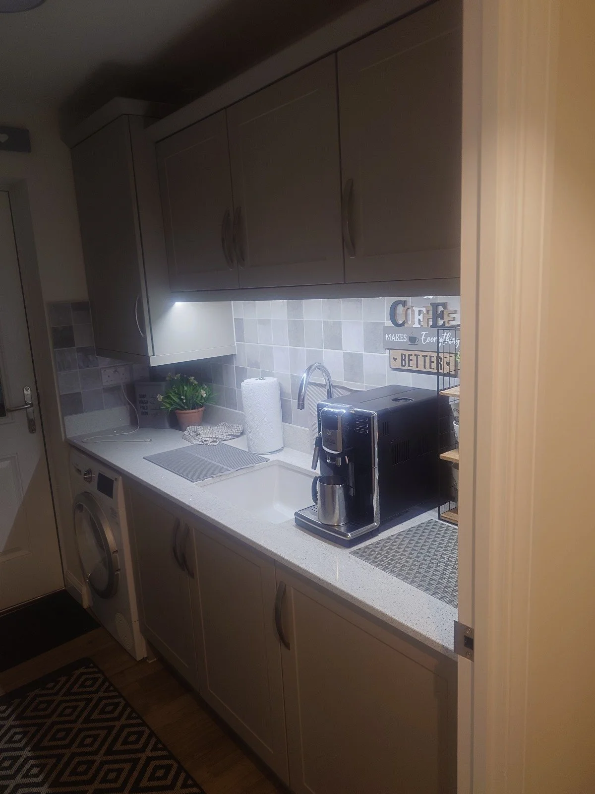 Modern kitchen with white cabinets, coffee machine on countertop, tiled backsplash, washing machine, and under-cabinet lighting.