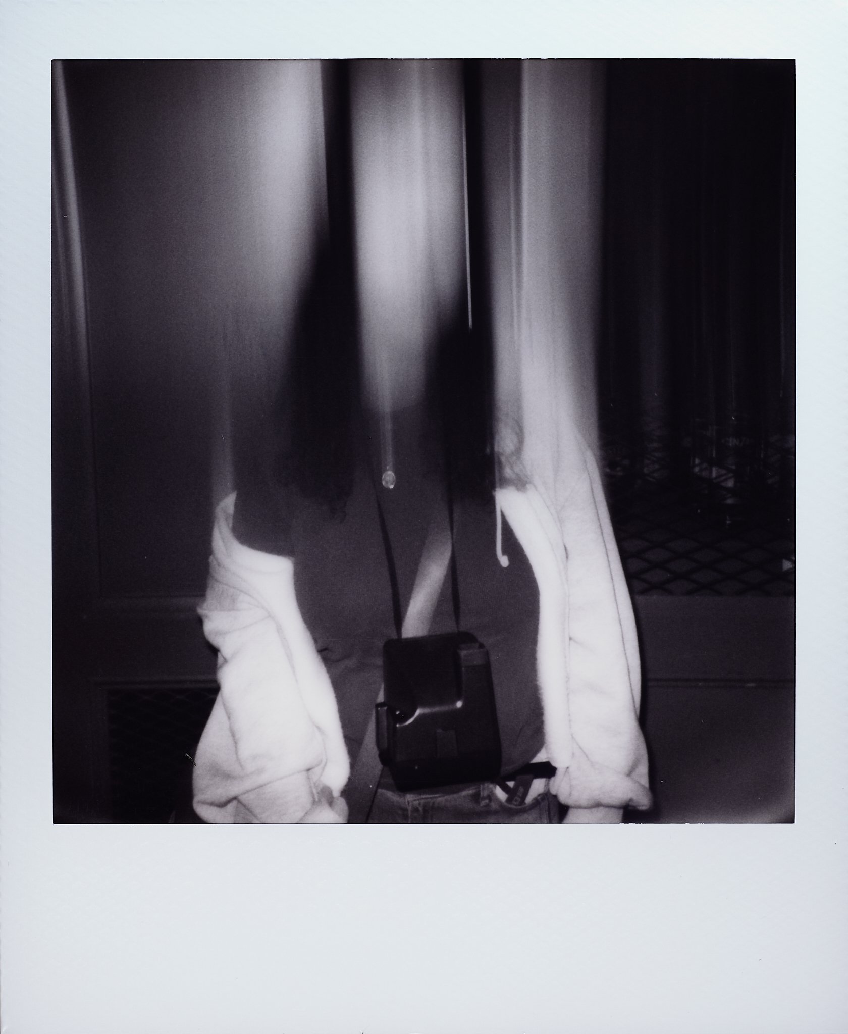 A person with long hair, wearing a hoodie and a backpack, standing in front of a dark door or wall.