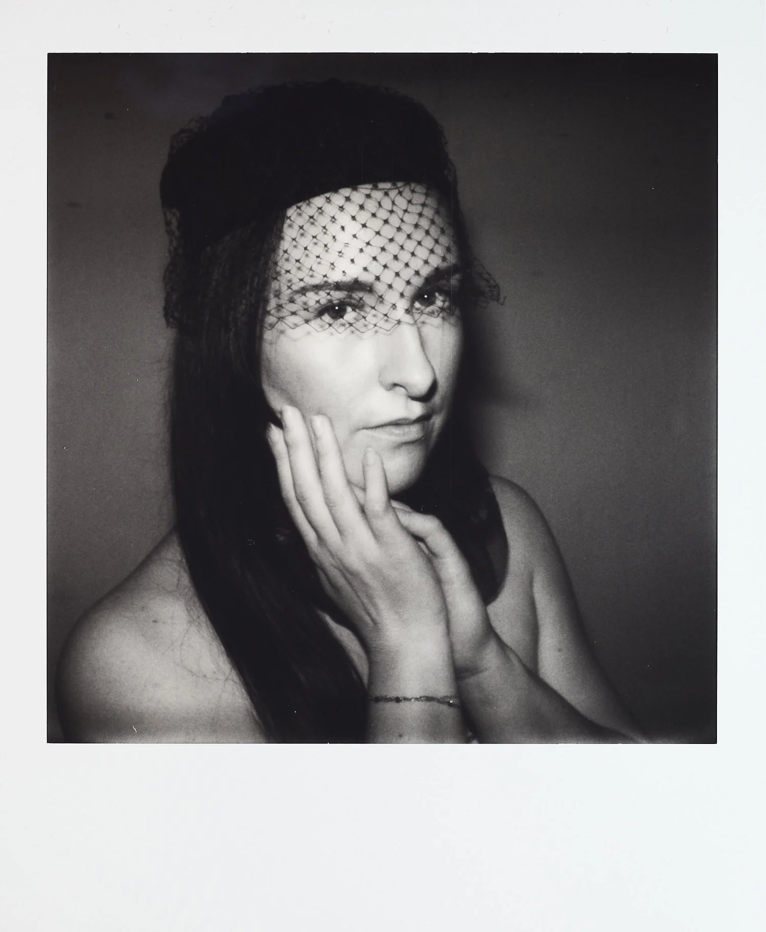 Black and white portrait of a woman with a netted veil covering part of her face, touching her cheek, with a neutral expression.