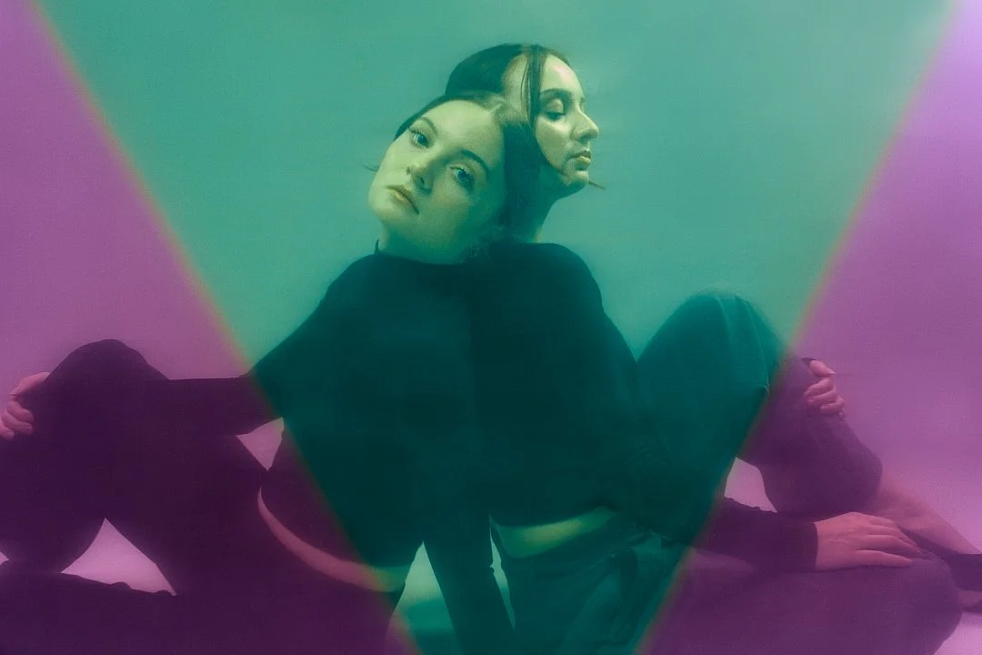 Two women sitting back-to-back with a mirrored effect on a pink and green gradient background.