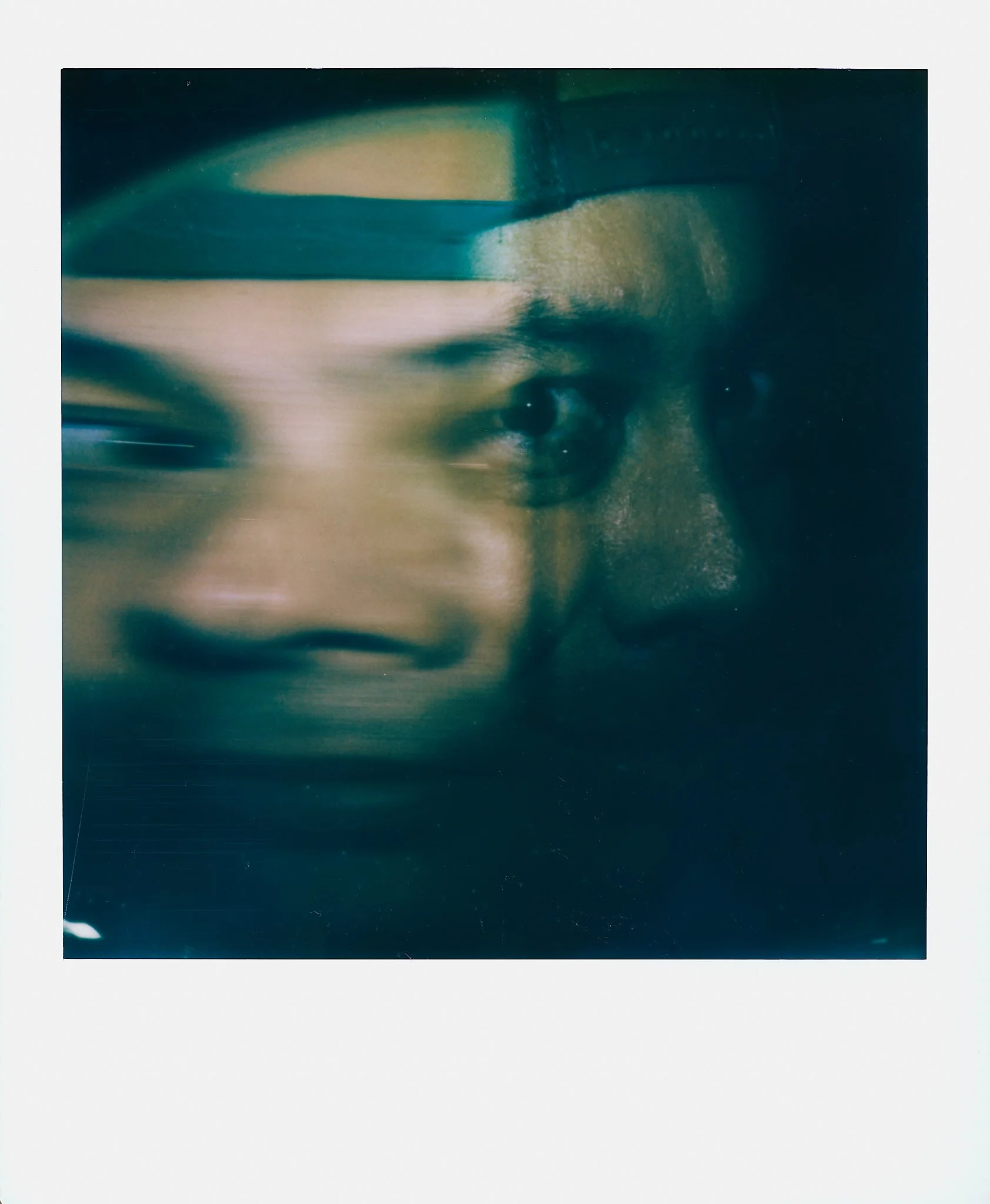 A close-up of two overlapping faces, one in focus with dark skin and the other blurred, wearing a hat, with a dark background.