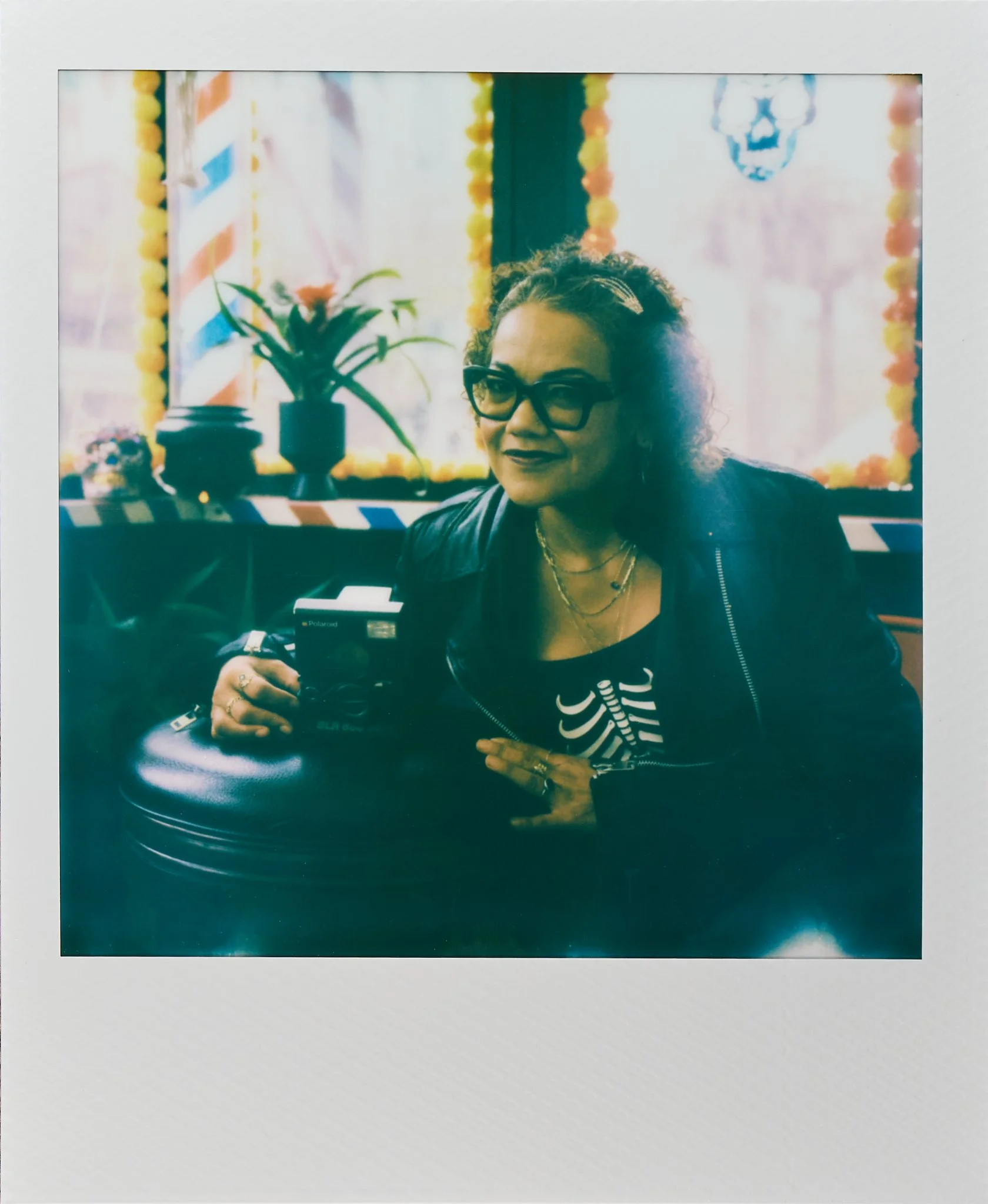 A woman with curly hair, glasses, and dark clothing sitting at a table in a decorated indoor setting with a plant and a decoration resembling a skull in the background.