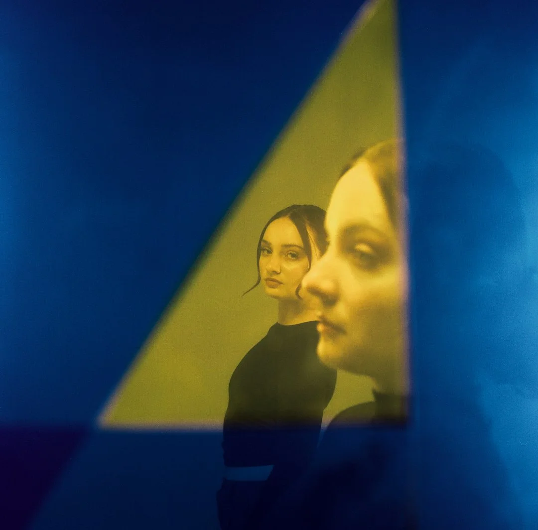 Two women are seen through a yellow triangular opening, with the background illuminated in blue. One woman looks at the camera, while the other is in profile, facing right.