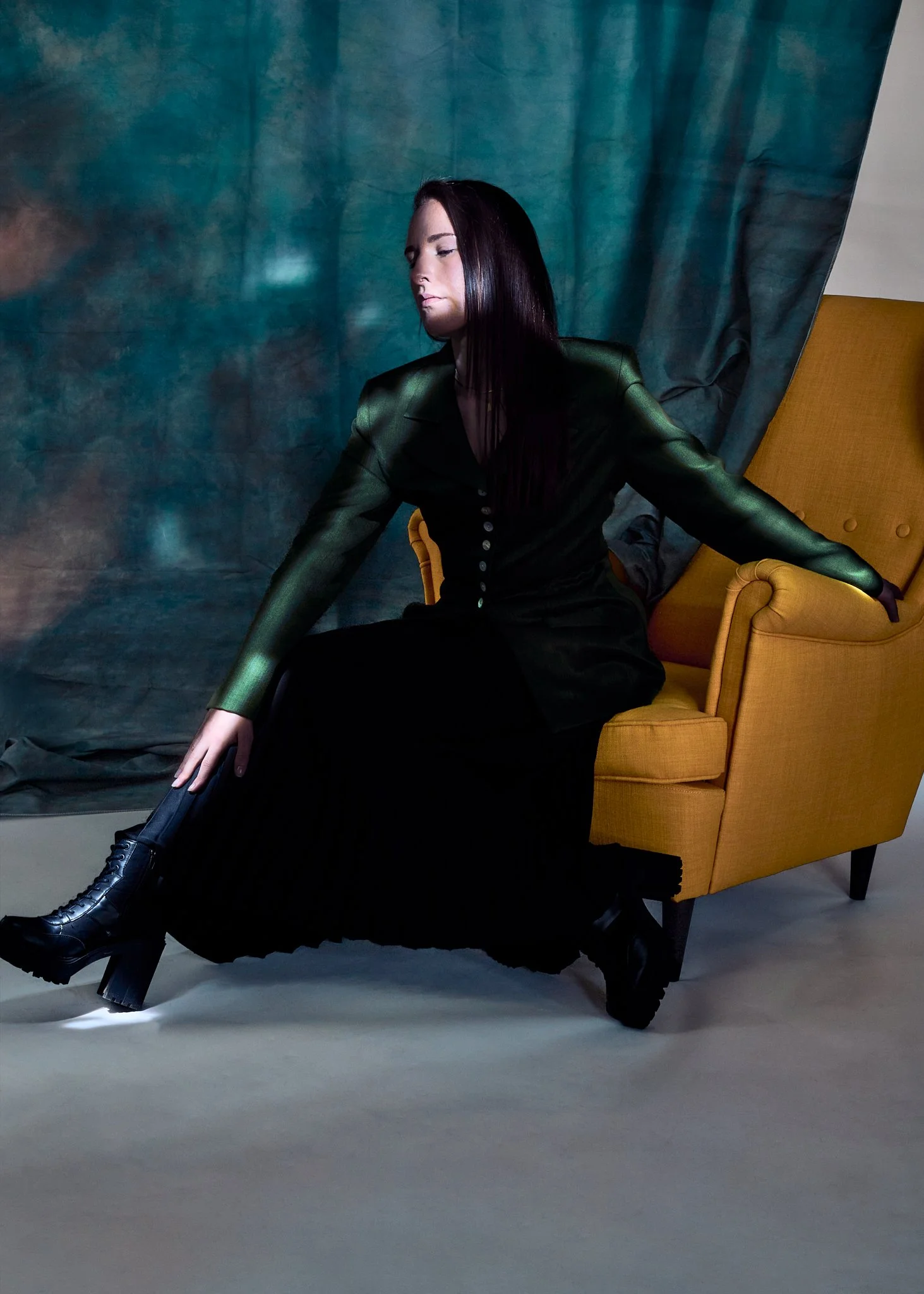 A woman with long dark hair, wearing a shiny green jacket and a long black skirt, sitting on a mustard yellow armchair with her eyes closed and head tilted slightly downward, in front of a blue and gray background.