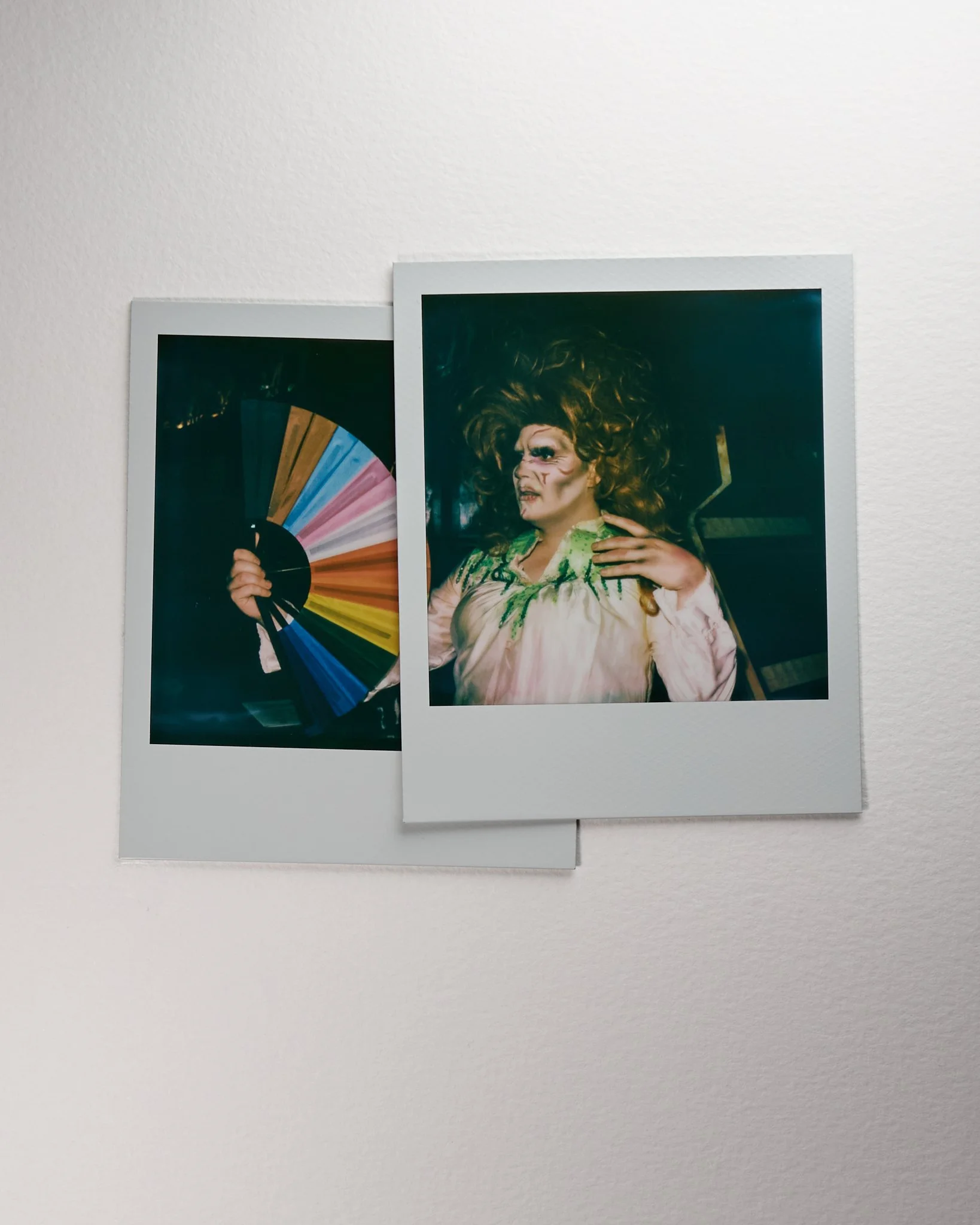 Two Polaroid photos on a white wall. One shows a person holding a colorful fan, the other shows a person in drag with elaborate makeup and a curly wig.