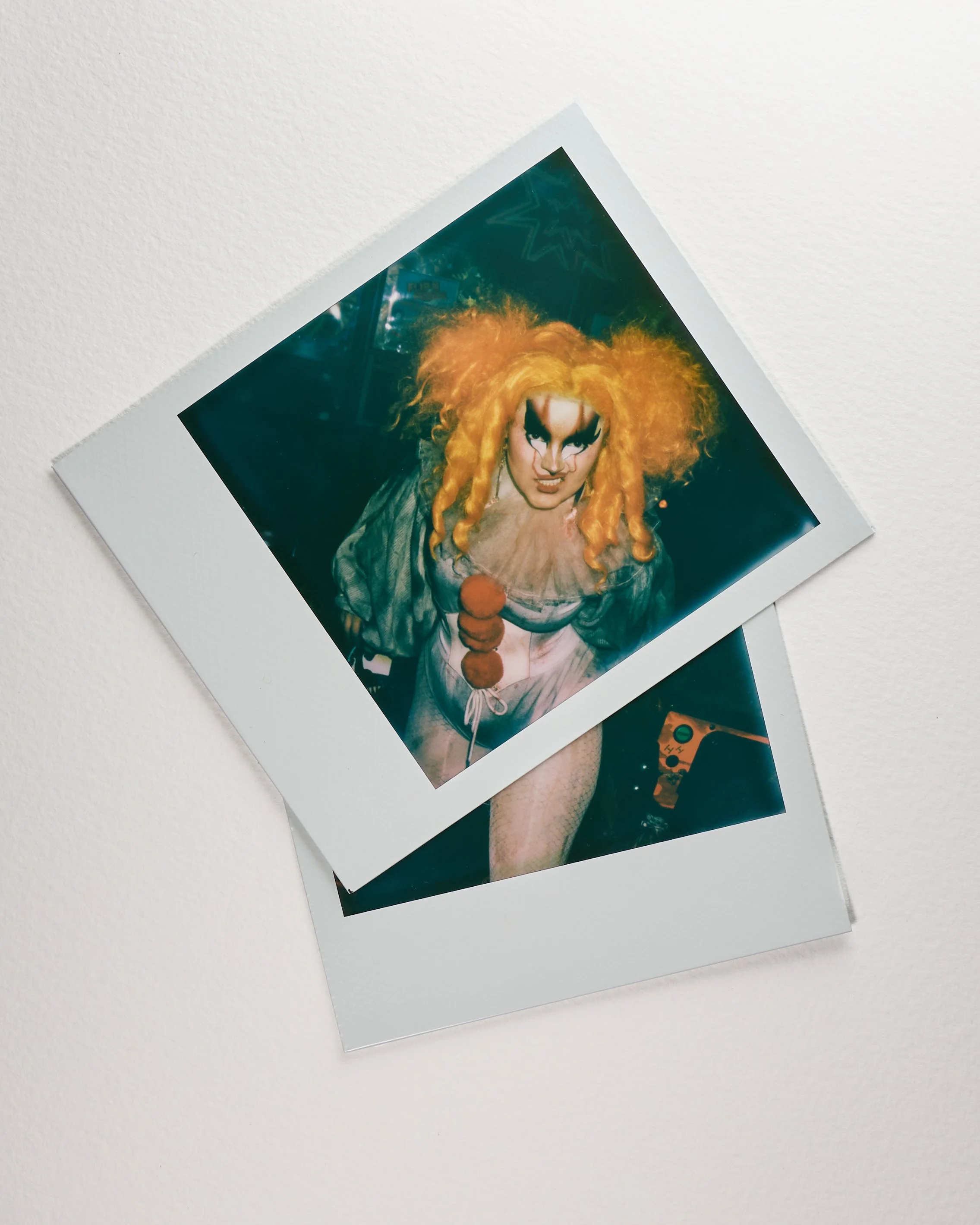 Two Polaroid photos on a white surface, one featuring a person with theatrical makeup and bright orange hair dressed as a clown or performer, and the other partially visible with some electronic equipment.