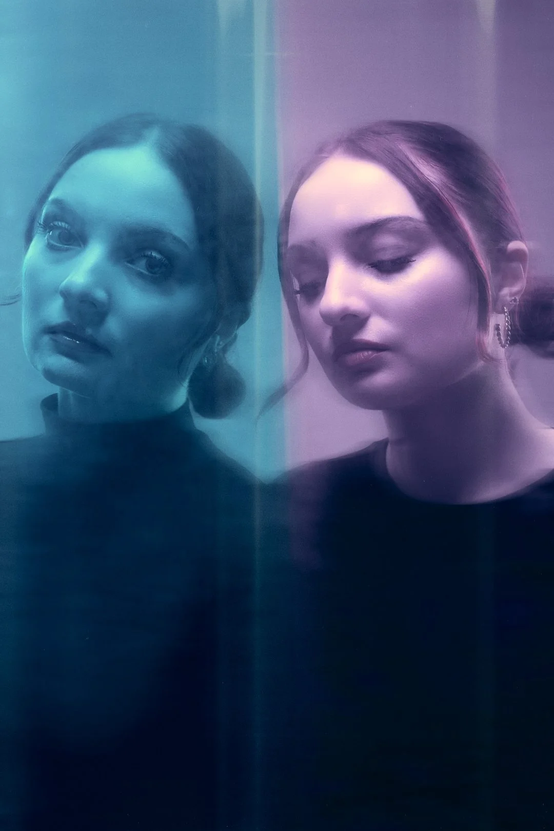 Two women with dark hair illuminated by blue and purple lighting, looking at the camera through a glass surface.