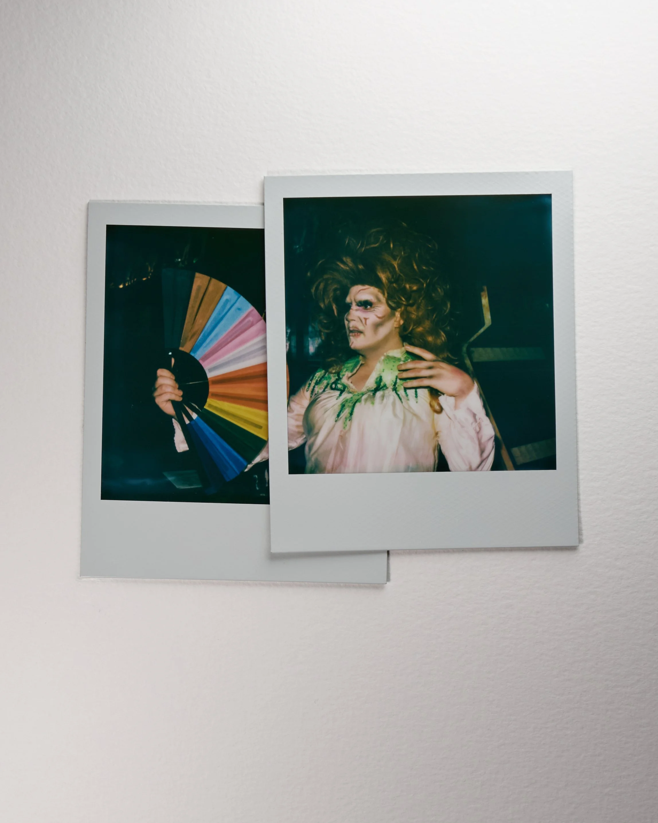 Two Polaroid photos on a white wall, one showing a person with elaborate makeup and costume, the other showing a colorful hand fan.