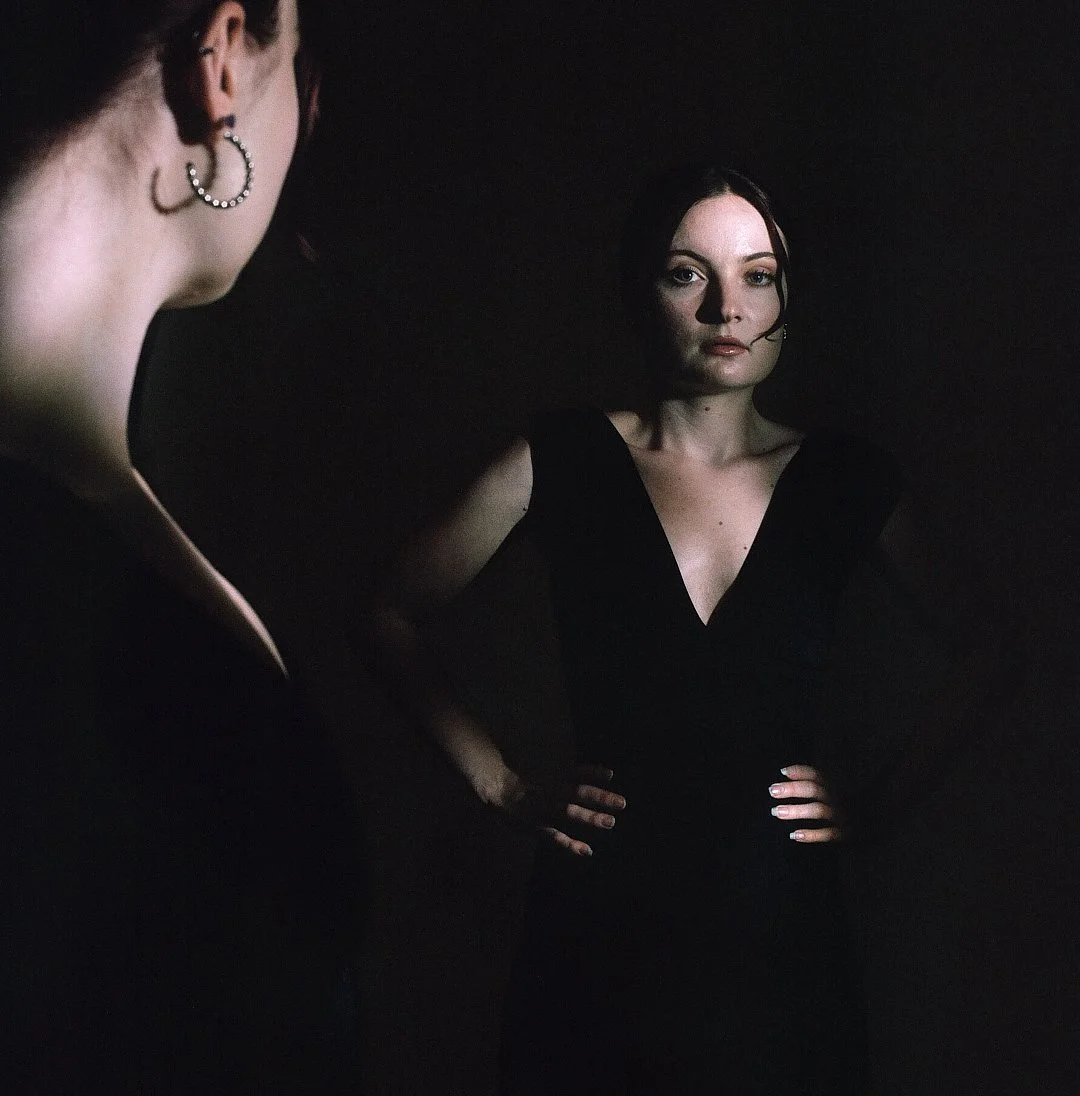 A woman with dark hair and a black top looking serious, standing in front of a mirror, with her reflection visible.