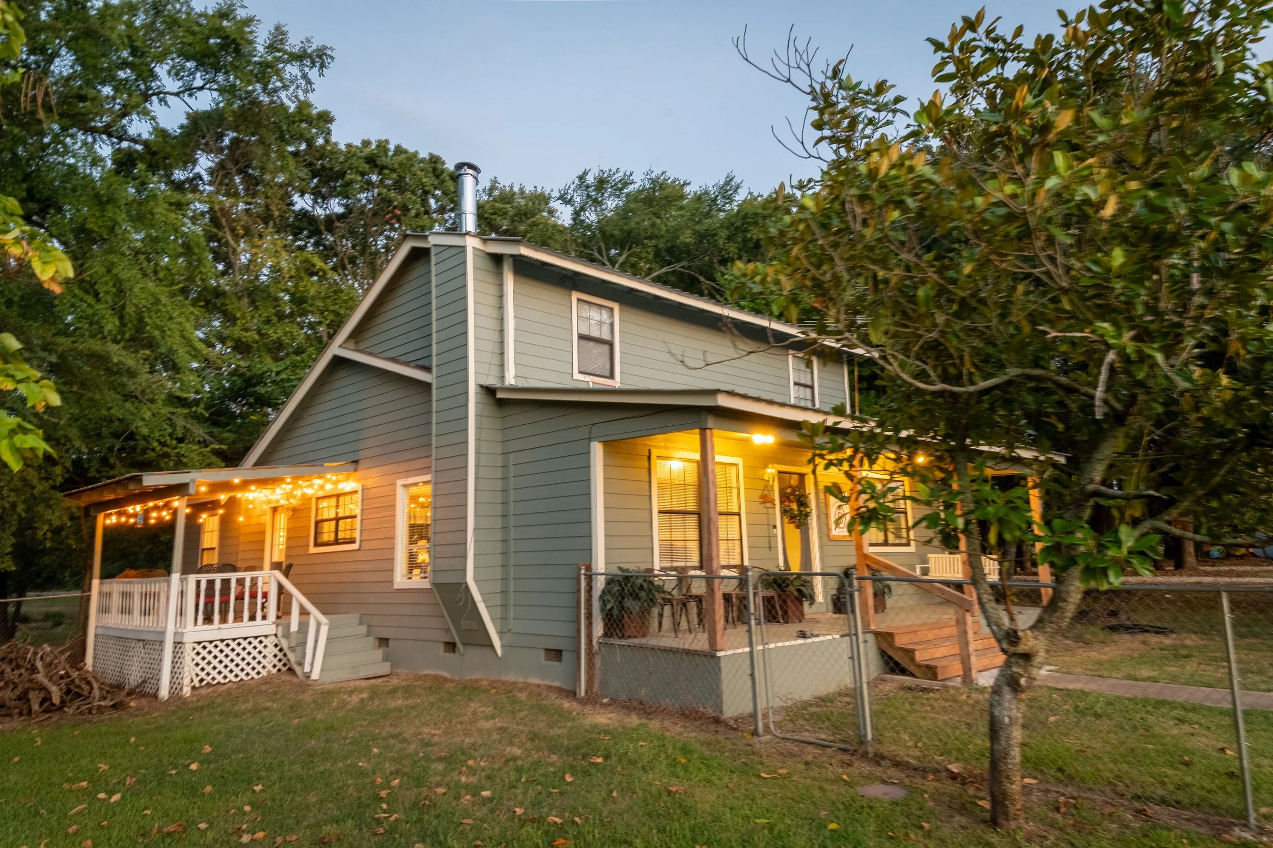 A two-story house with green siding and a small front porch, decorated with string lights and warm interior lighting, surrounded by trees and a grassy yard with a chain-link fence.