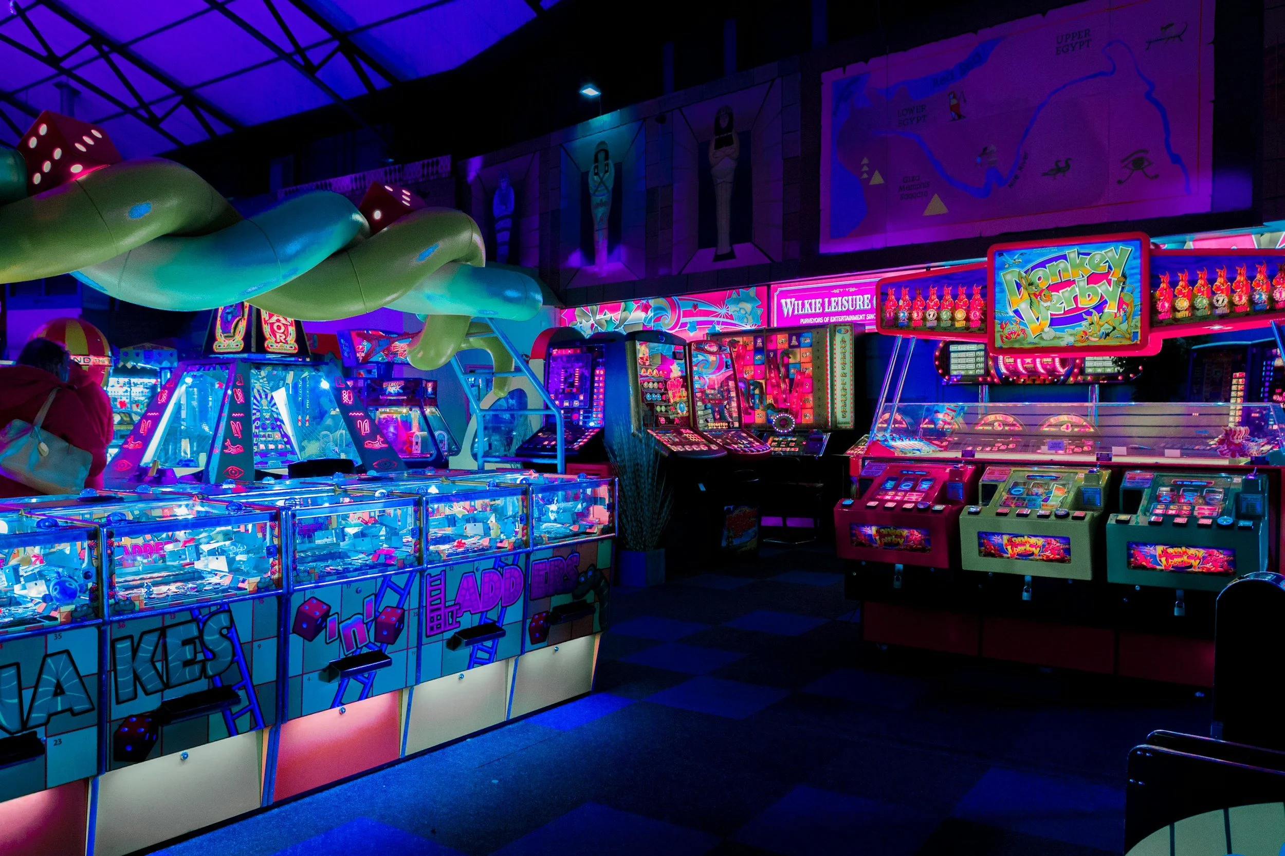 Arcade with neon lighting, featuring various gaming machines including pinball and claw machines, and colorful inflatable slides overhead.
