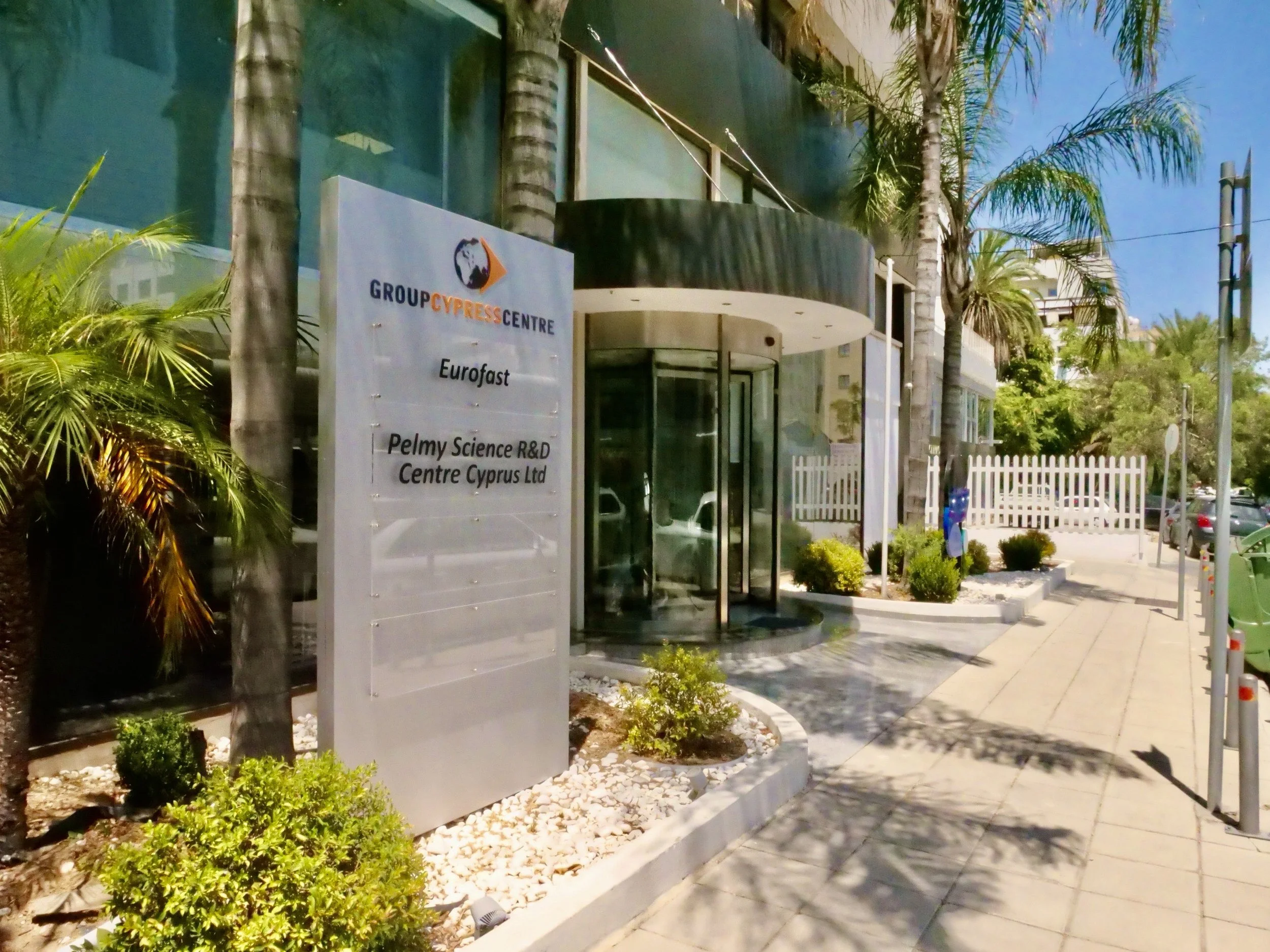 Sign outside the Group Cypress Centre for Eurofast and Pelmy Science R&D Centre Cyprus Ltd, with palm trees, plants, and a building entrance in the background.