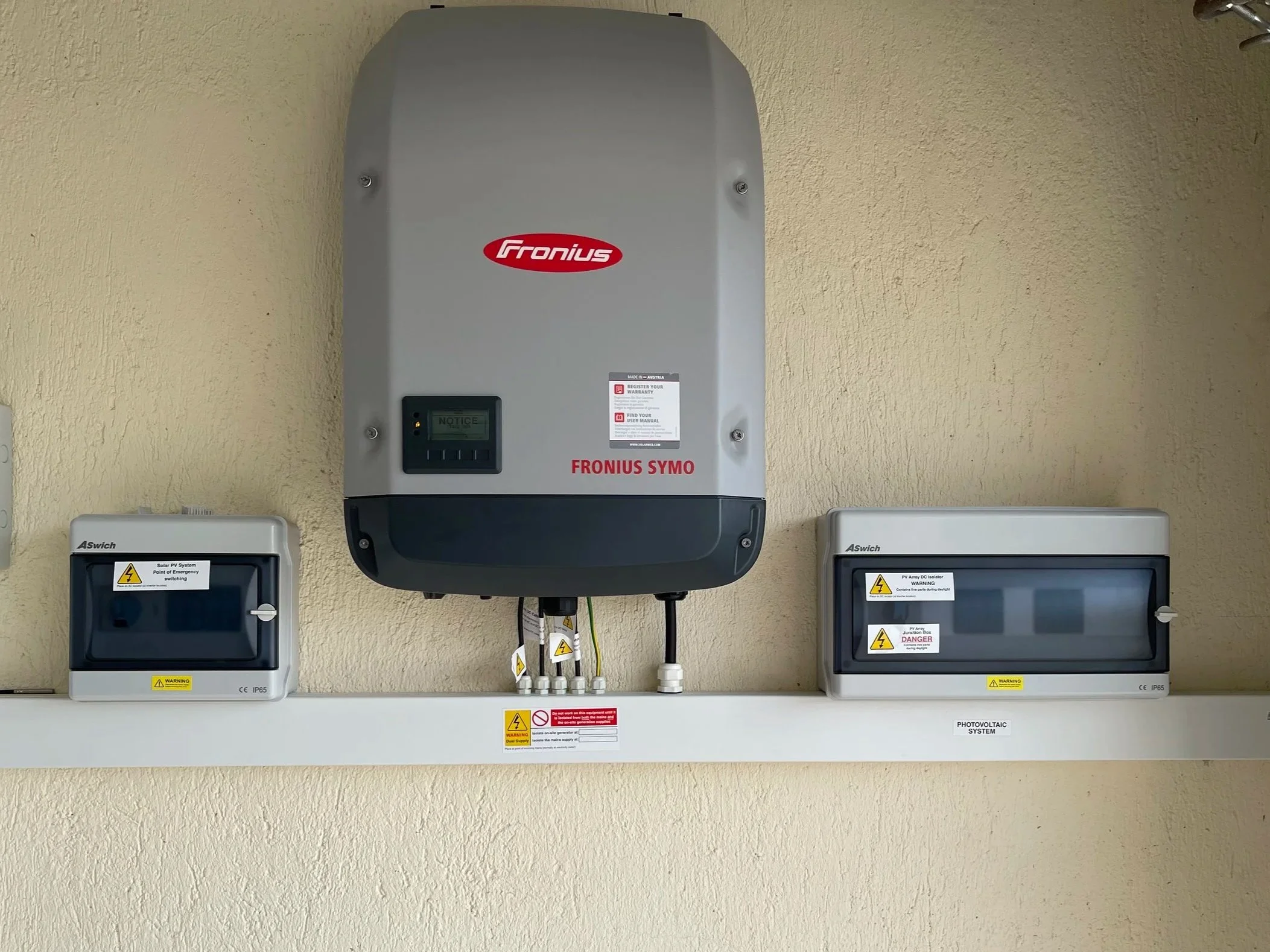 An outdoor electrical installation with a large gray solar inverter labeled 'Fronius SYMO' mounted on a beige wall. Below the inverter are two gray electrical boxes with warning labels, and various cables are connected to the system, indicating it is part of a photovoltaic solar power setup.
