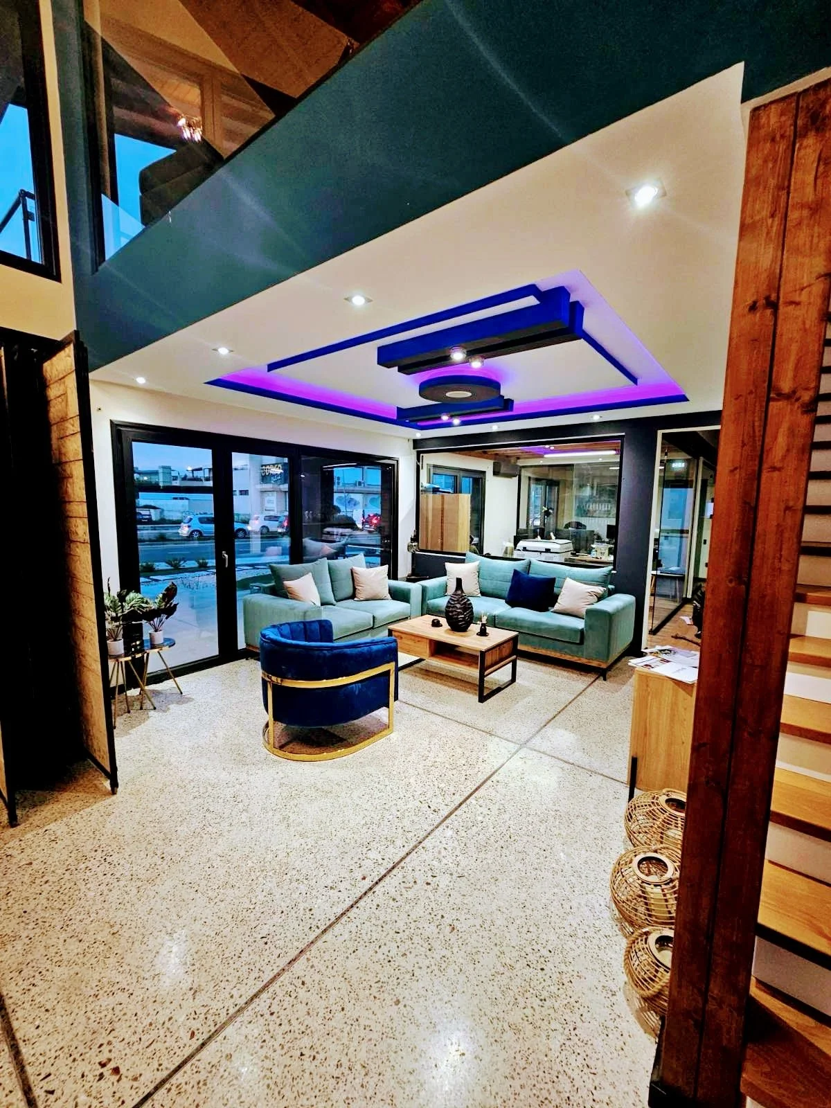Interior of a modern living room with large glass sliding doors, a green sofa with pillows, a blue velvet armchair, a wooden coffee table with decorative items, and a black and purple ceiling feature with built-in lighting.