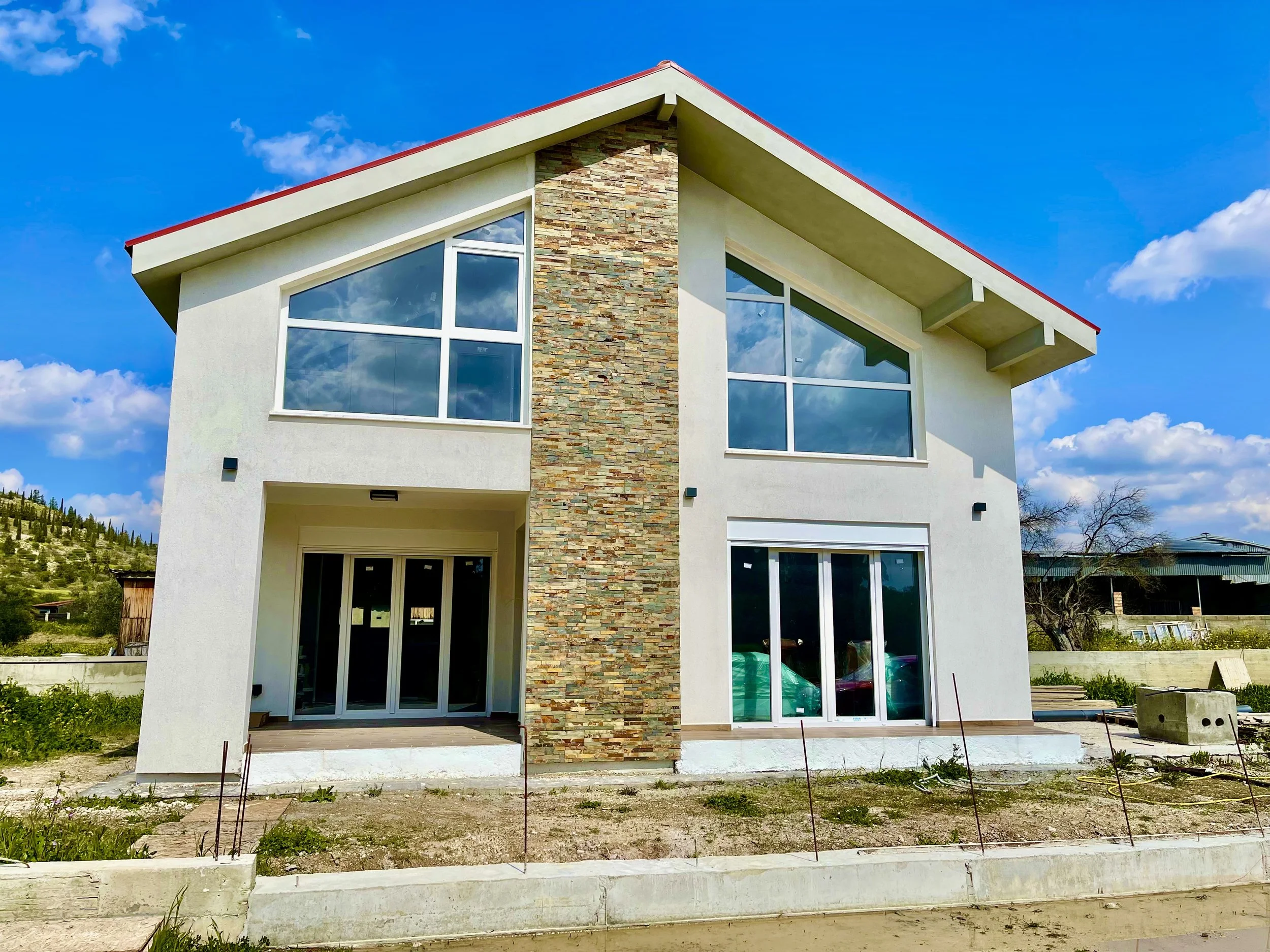 Modern two-story house under construction with large windows, a stone accent wall, and a pitched roof, set against a blue sky with some clouds.