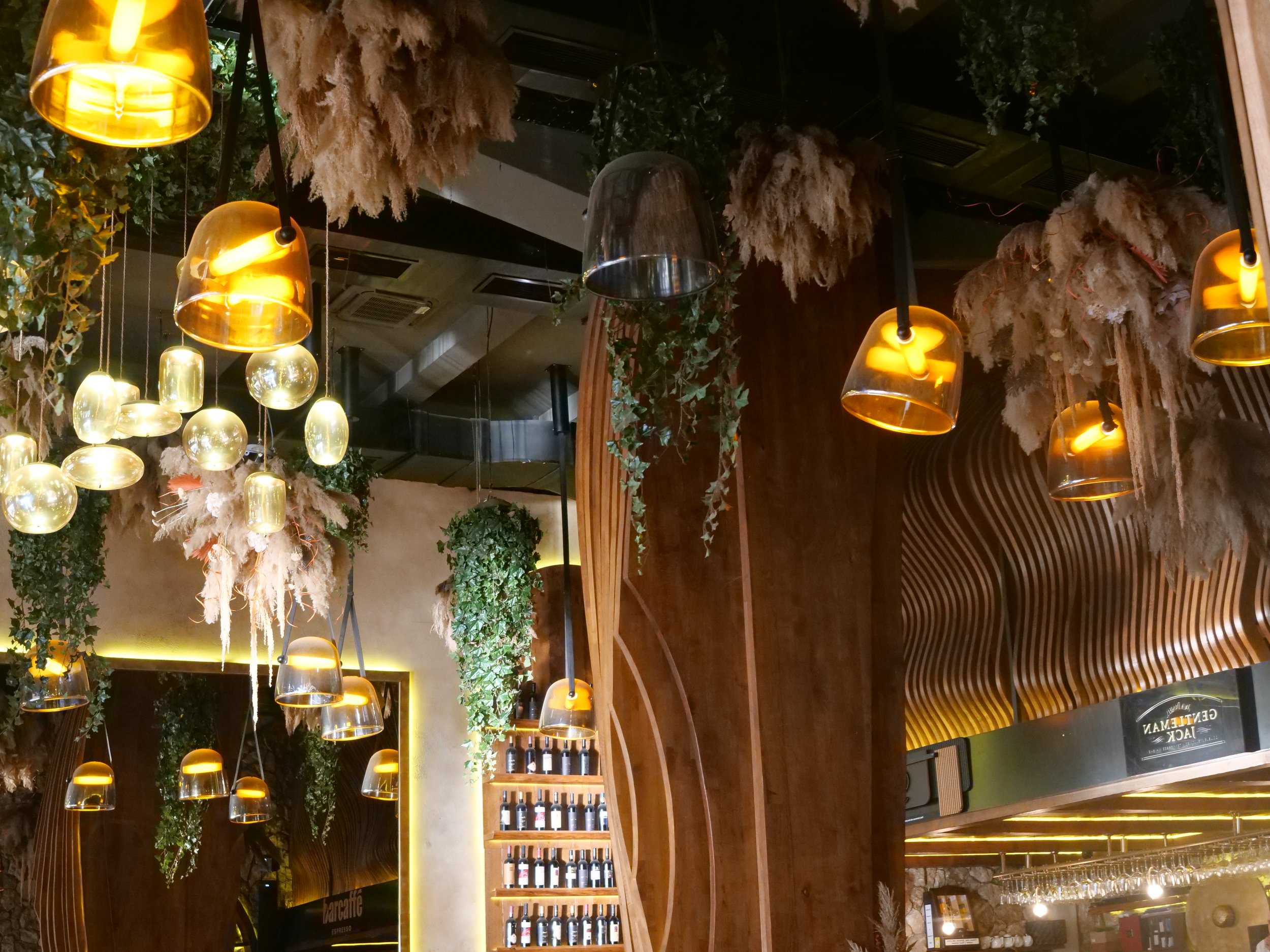 Interior of a restaurant or cafe with hanging amber-colored pendant lights, greenery, and decorative dried plants.