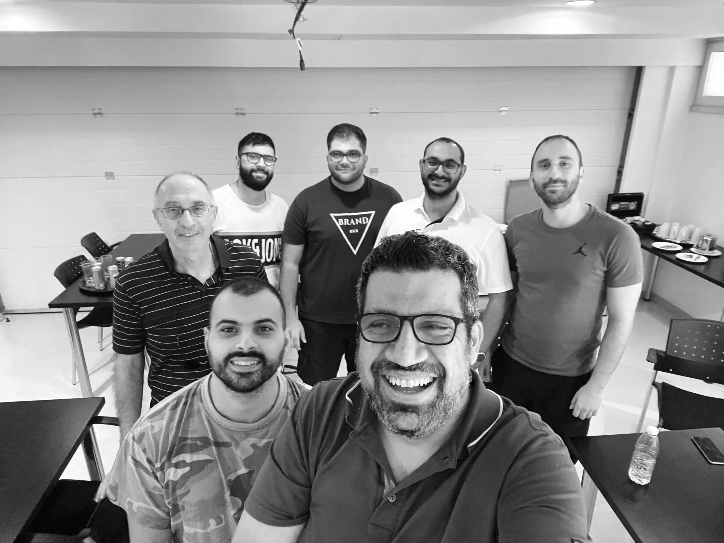 A group of six men posing for a photo in an indoor setting, with tables and drinks in the background.