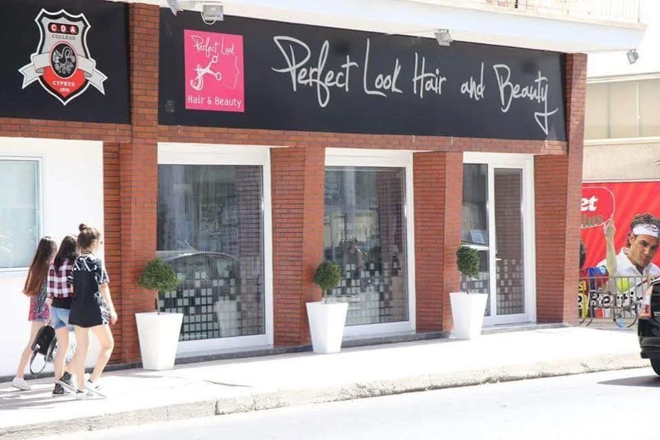Three young women walking past a hair and beauty salon with large windows, potted plants outside, and signage that reads "Perfect Look Hair and Beauty" on a black background. A partial view of a person in a colorful jersey and a vehicle can also be seen.