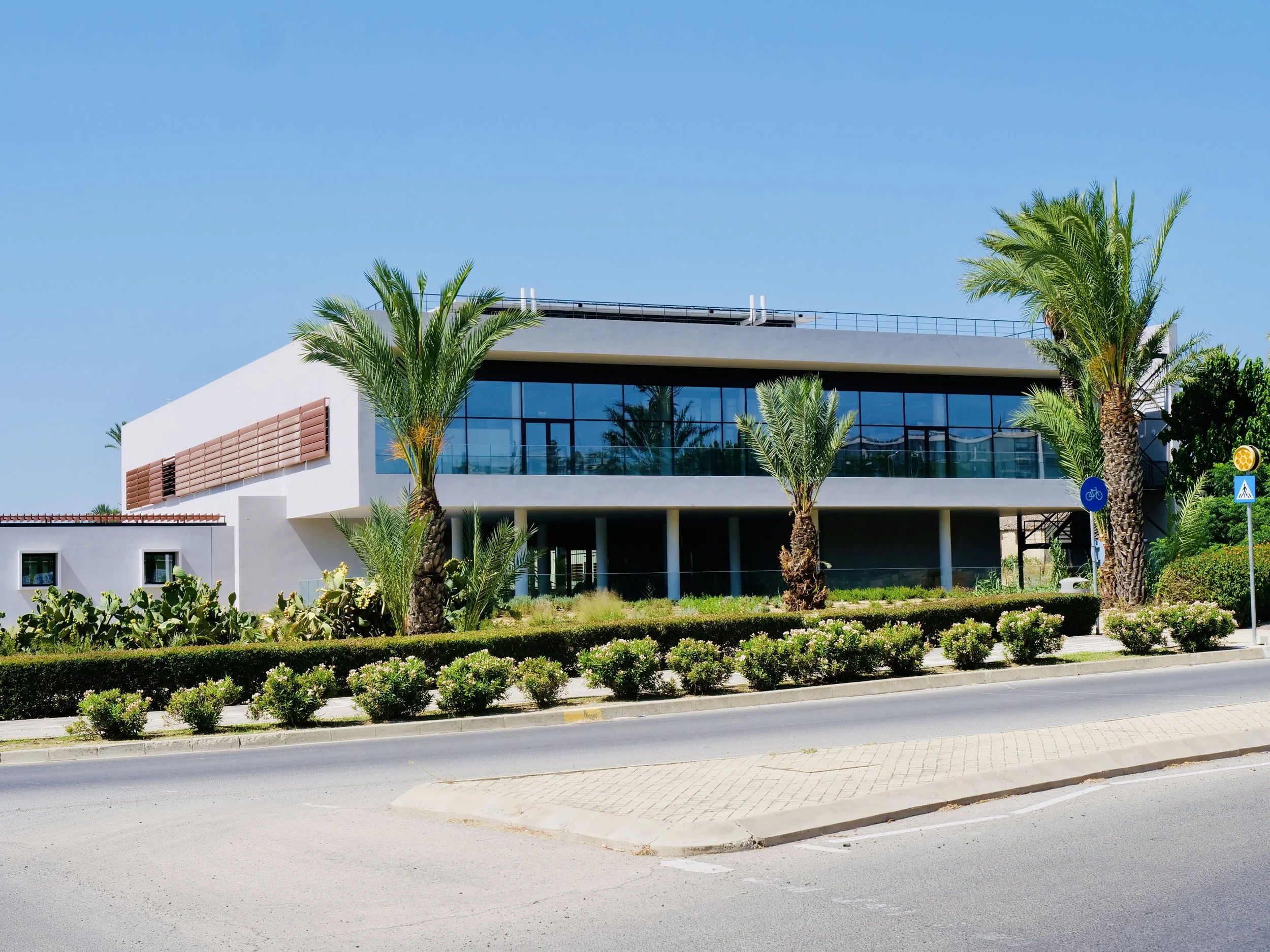Modern building with glass windows, surrounded by palm trees and landscaped bushes, under a clear blue sky.