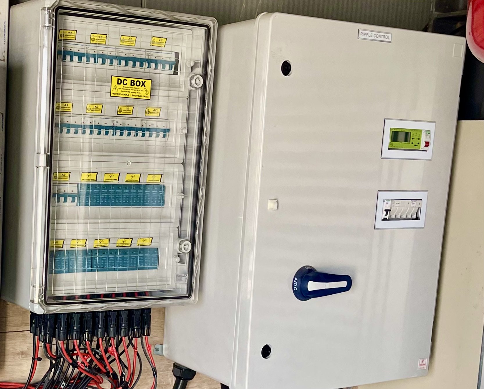 Electrical control panel with a transparent cover, yellow warning labels, blue circuit breakers, and red and black wires connected to a large white box labeled 'Ripple Control'.