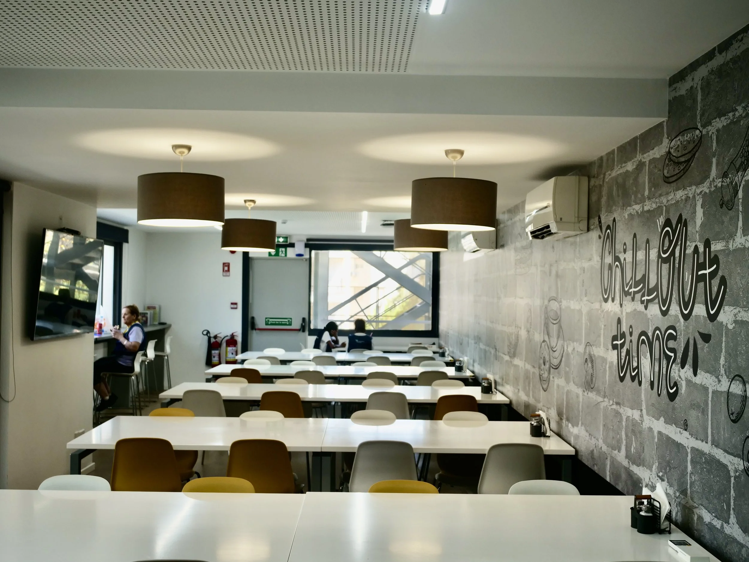 Interior of a modern cafeteria or dining area with white tables, chairs in various colors, large pendant lights, a wall with decorative drawings of food items and words, large windows allowing natural light, and ceiling-mounted air conditioning units.