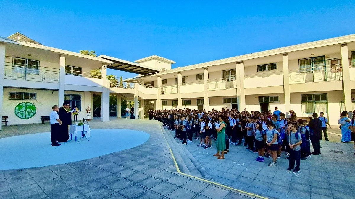 Students gathered outdoors in school uniform, standing in organized rows, listening to a speaker at a ceremony in a school courtyard on a sunny day.