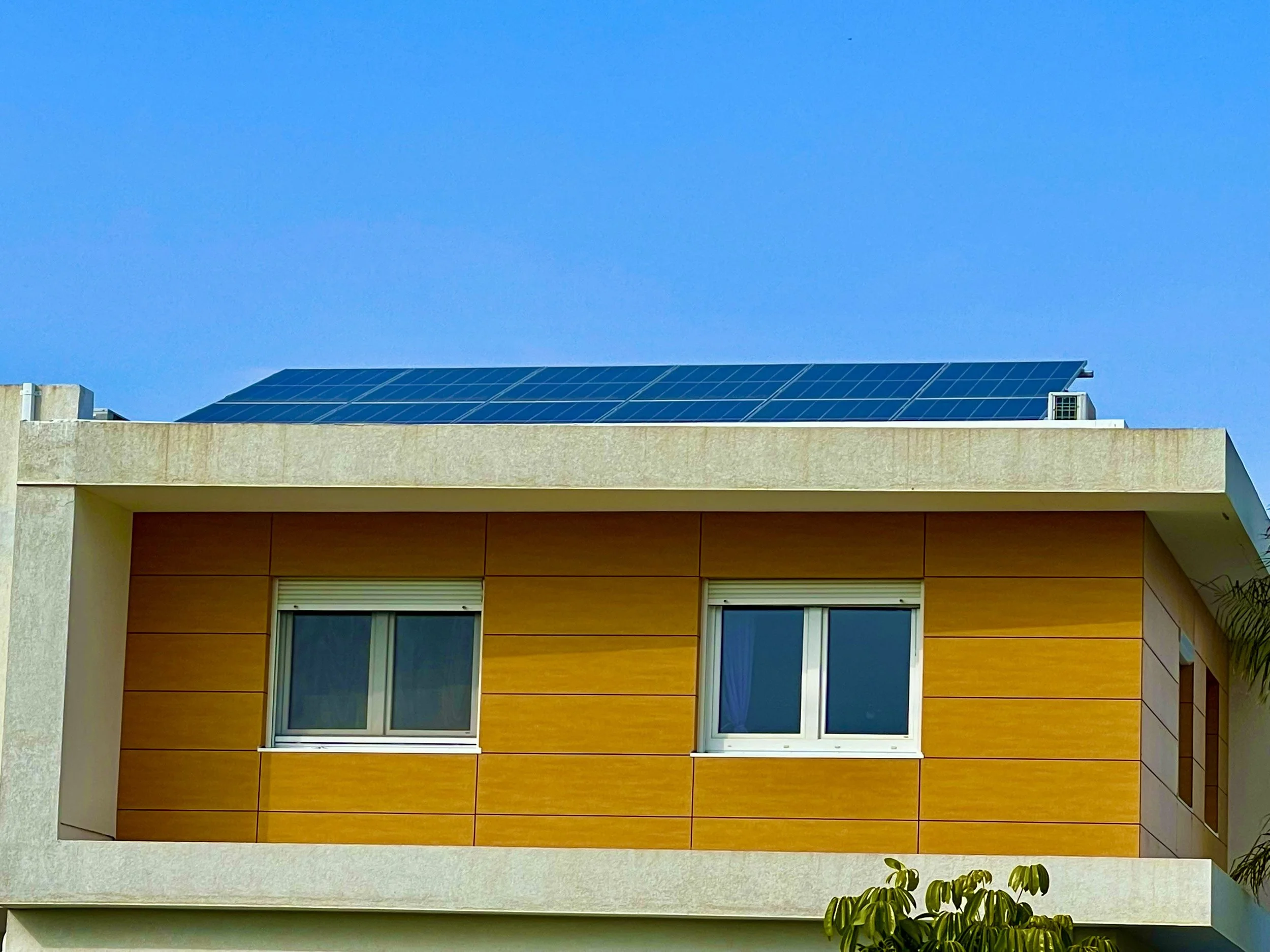 A modern building with solar panels on the roof, a beige facade, and large windows against a clear blue sky.