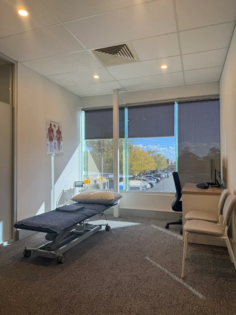Empty medical examination room with a hospital bed, pillow, and a cart near the window. Large window with blinds showing a parking lot and trees outside. Desk with a computer and several chairs.