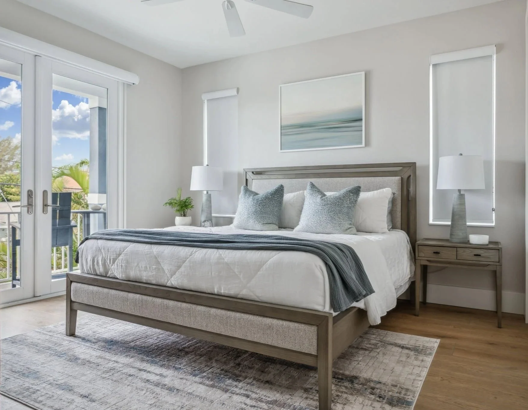 Modern bedroom with a king-size bed, white bedding, and blue pillows, flanked by two bedside tables with lamps. Large window with blinds revealing an outdoor view. Minimalist artwork on the wall above the bed and a light gray area rug on wooden floor