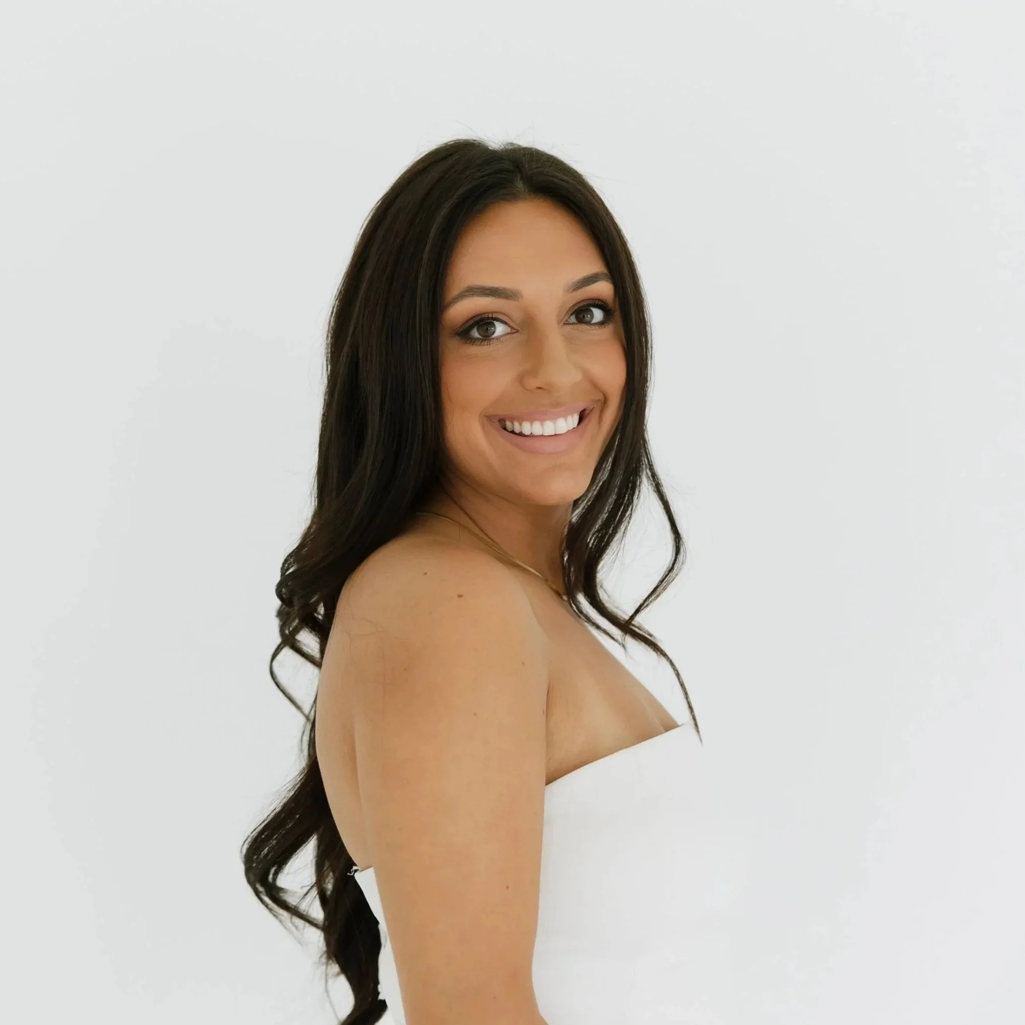 Smiling woman with long dark hair, tan skin, and white strapless top, standing against a plain white background.