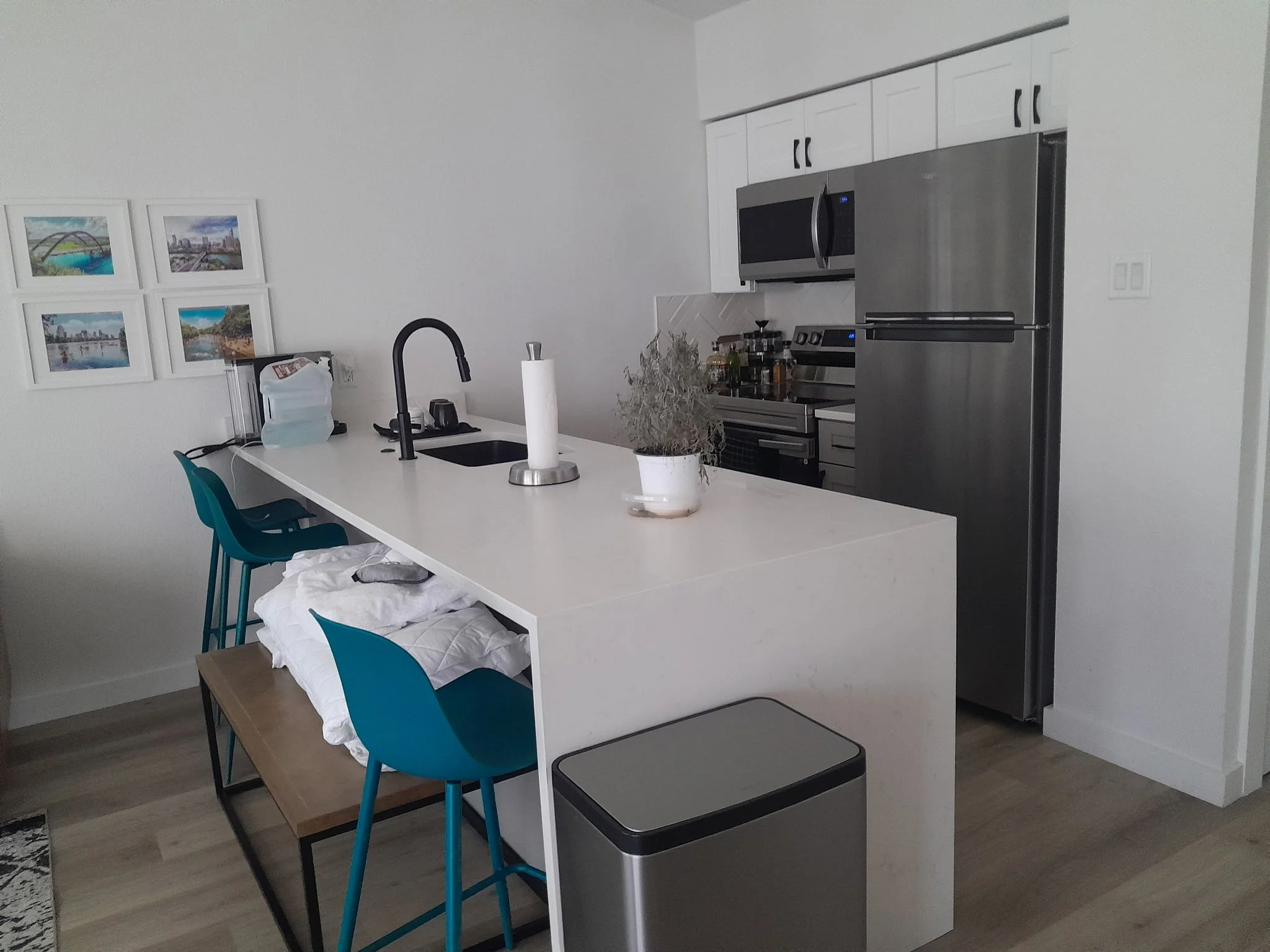 A modern kitchen with a white island, blue chairs, stainless steel refrigerator, microwave, and stove, with framed cityscape and nature photographs on the wall, and a small potted plant on the island.