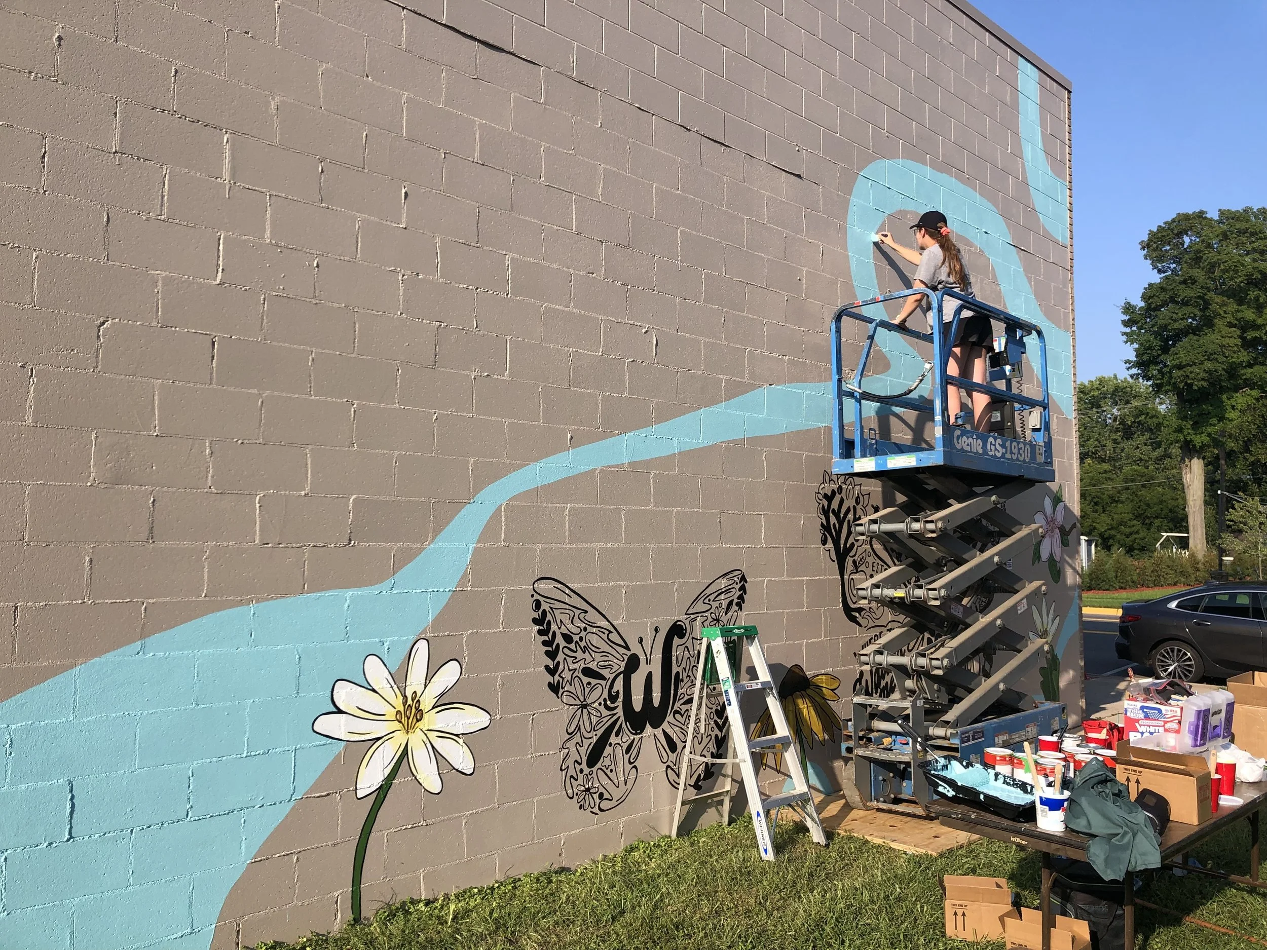 A woman on a blue lift painting a mural of a blue and black butterfly, a white flower, and a pink flower on a brick wall outside during the daytime.