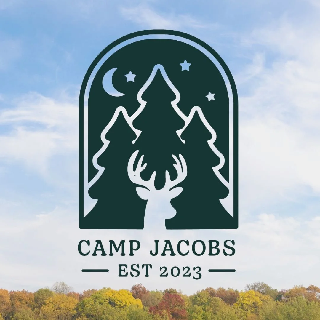 A logo featuring a deer with large antlers, surrounded by three pine trees, a moon, and stars. Text below reads "CAMP JACOBS EST 2023." The background shows a sky with clouds and a treeline with fall foliage.