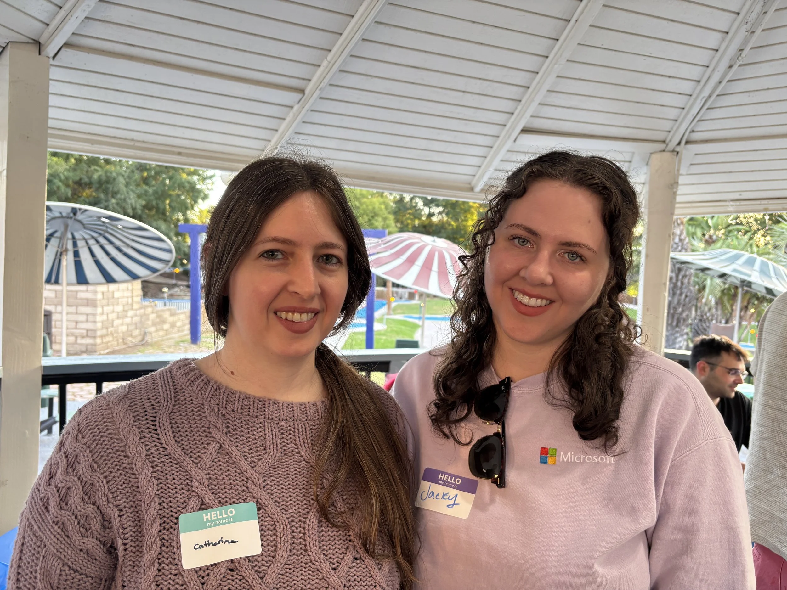 Two women standing together outdoors under a white roof, smiling at the camera. The woman on the left has long brown hair, wearing a purple knitted sweater, with a name tag reading 'Catherine.' The woman on the right has curly brown hair, wearing a l