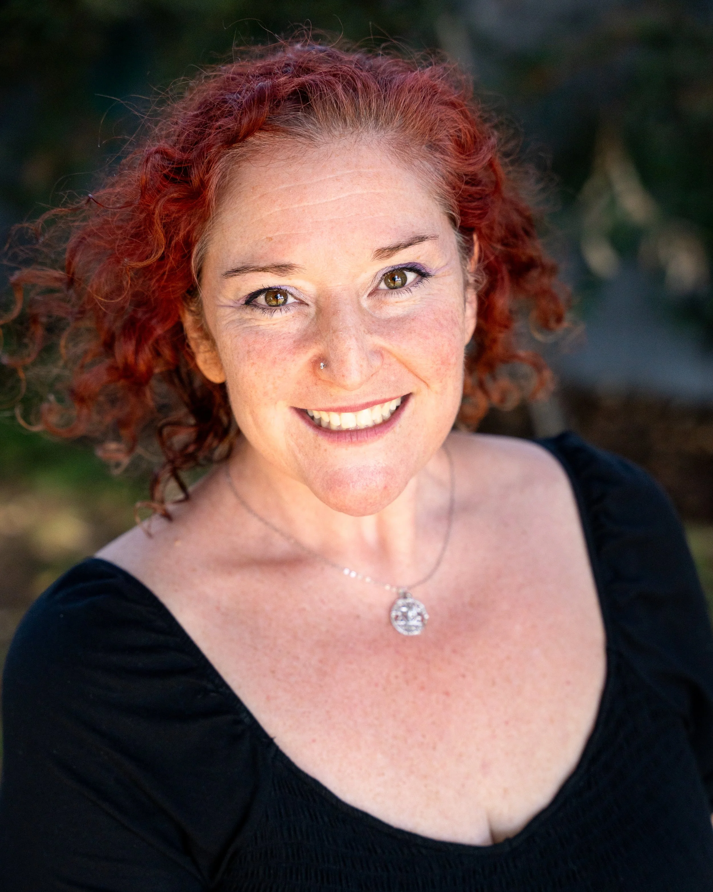 A woman with dark red hair smiling outdoors on a porch, wearing a turquoise dress with a bow at the neckline and a beige cardigan.
