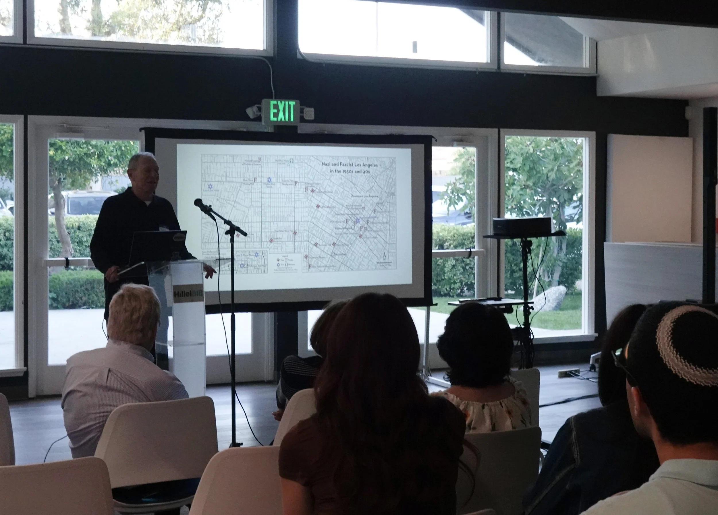 A man presents a lecture in a room with large windows, a projector screen displaying a map of Los Angeles during the 1930s and 1940s, and an audience seated facing him.