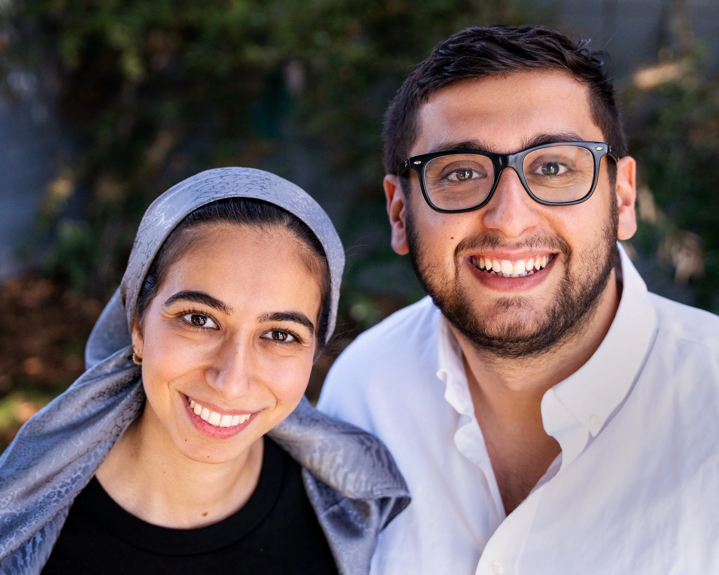 A woman and man smiling for a selfie indoors at night, with a window showing lights outside.