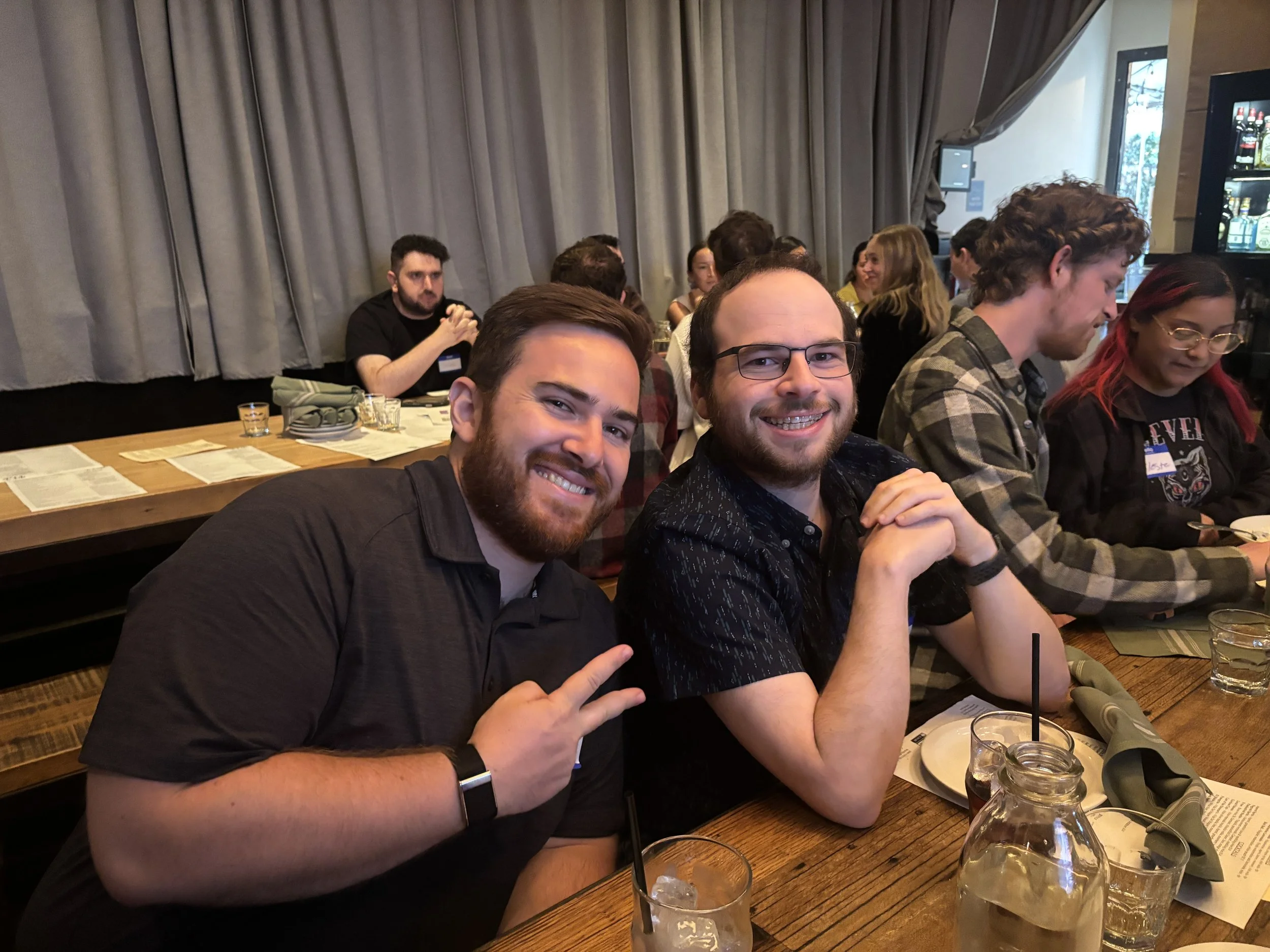 A group of people gathered around a table at a restaurant, with some smiling and making peace signs, while others are engaged in conversation.