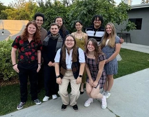Group of nine young adults standing outdoors in front of a house and greenery, smiling for a photo.