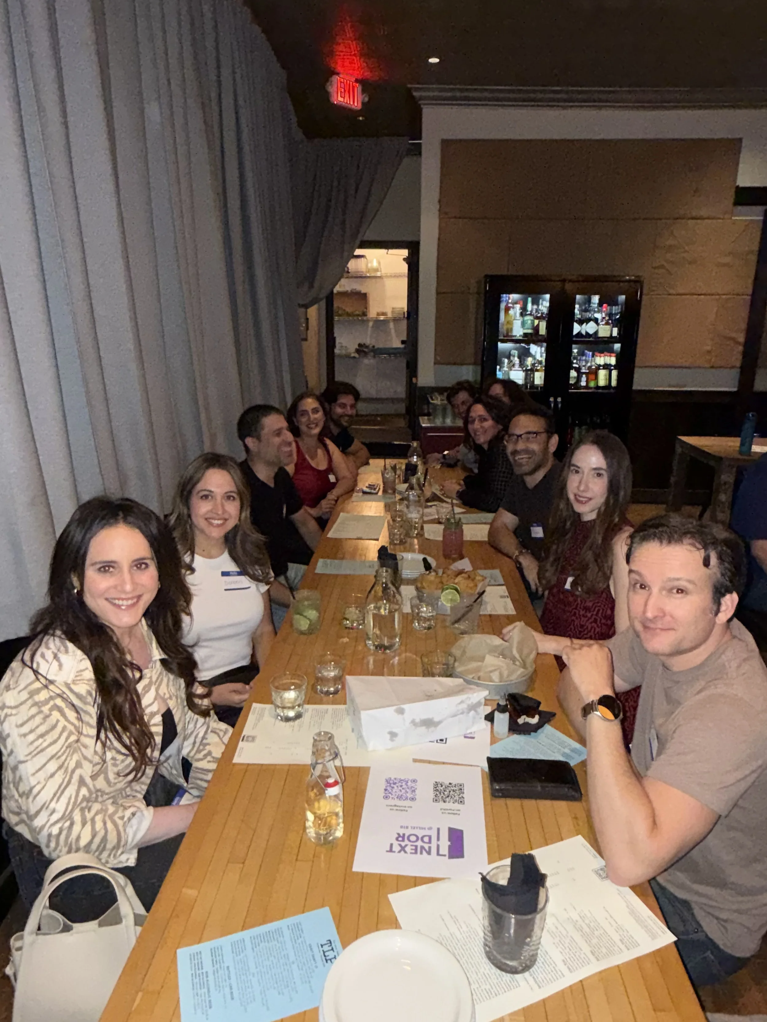 Group of people sitting around a long table in a restaurant, smiling and enjoying food and drinks, with menus, water glasses, and some snacks visible on the table.
