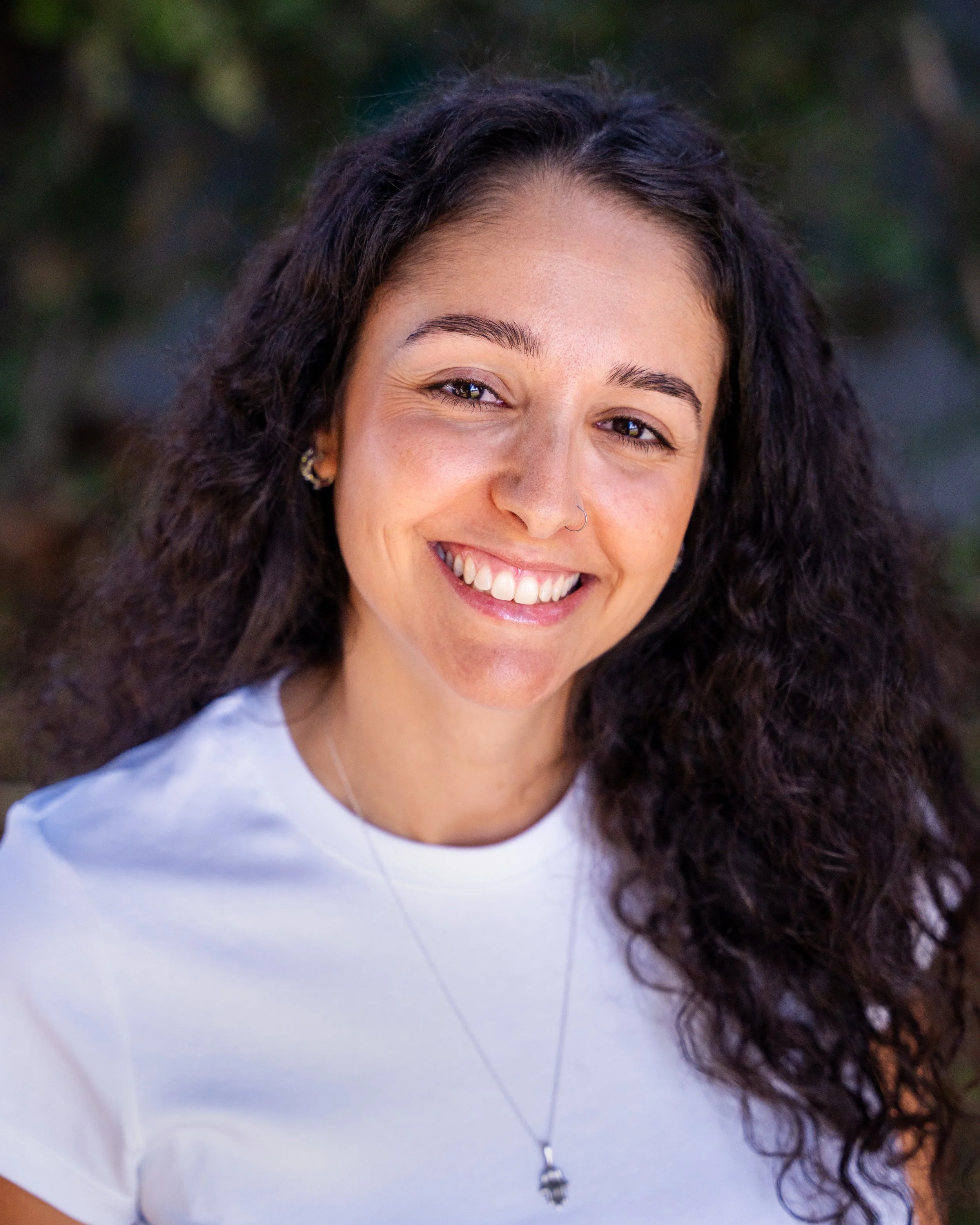 A young woman with curly black hair, smiling, wearing a white sleeveless top with ruffled straps, in front of a blurred outdoor background.