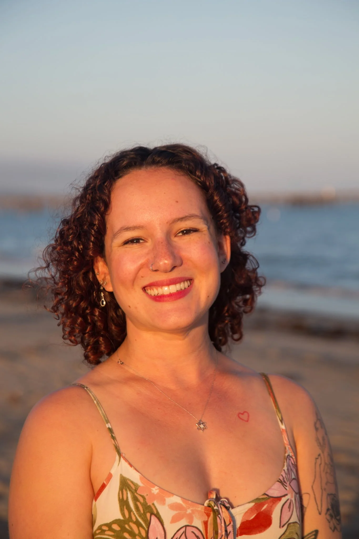 A smiling woman with curly hair standing on a beach during sunset.