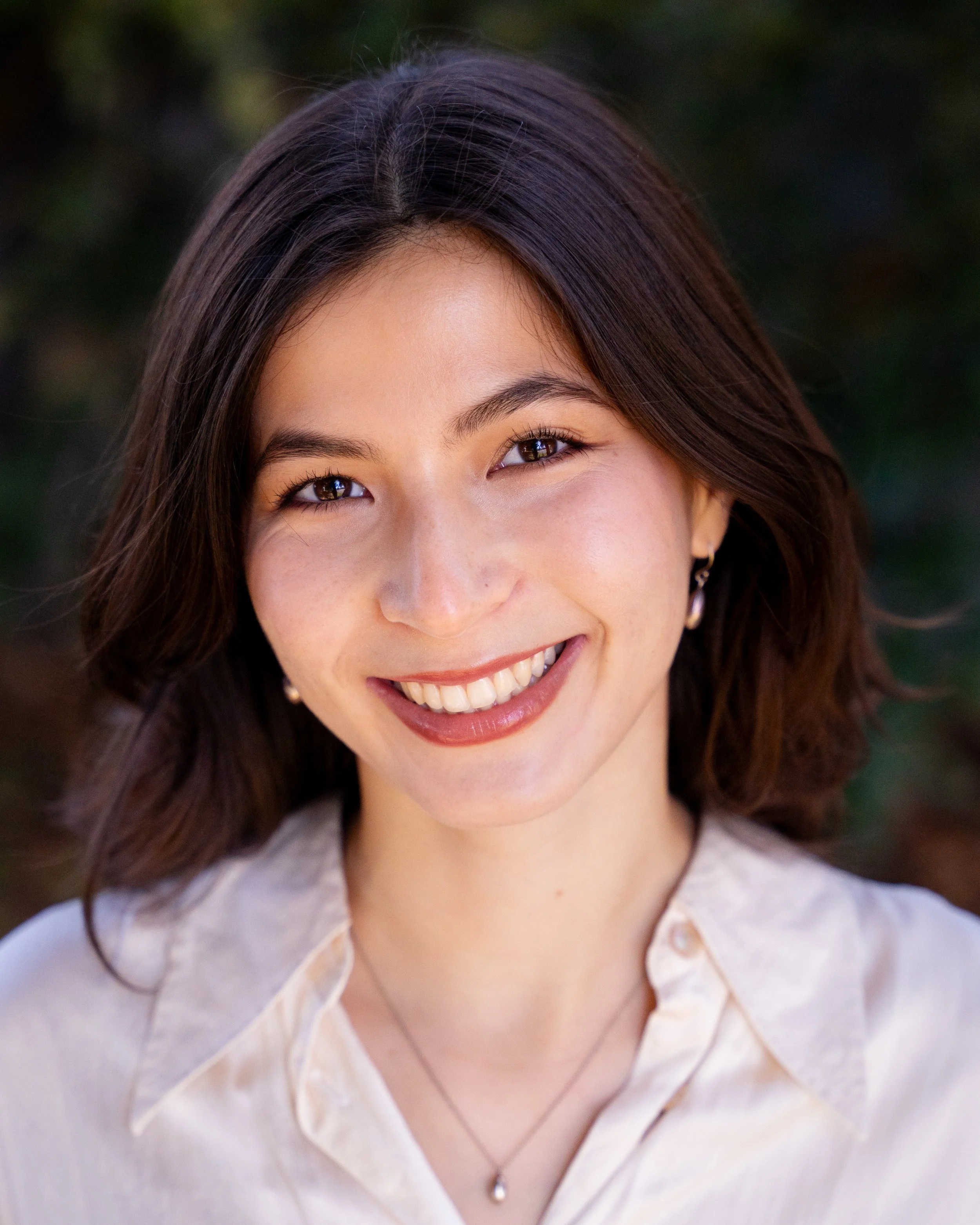 A young Asian woman with long black hair, wearing a black top and a delicate necklace, smiling softly in front of a plain light-colored wall.