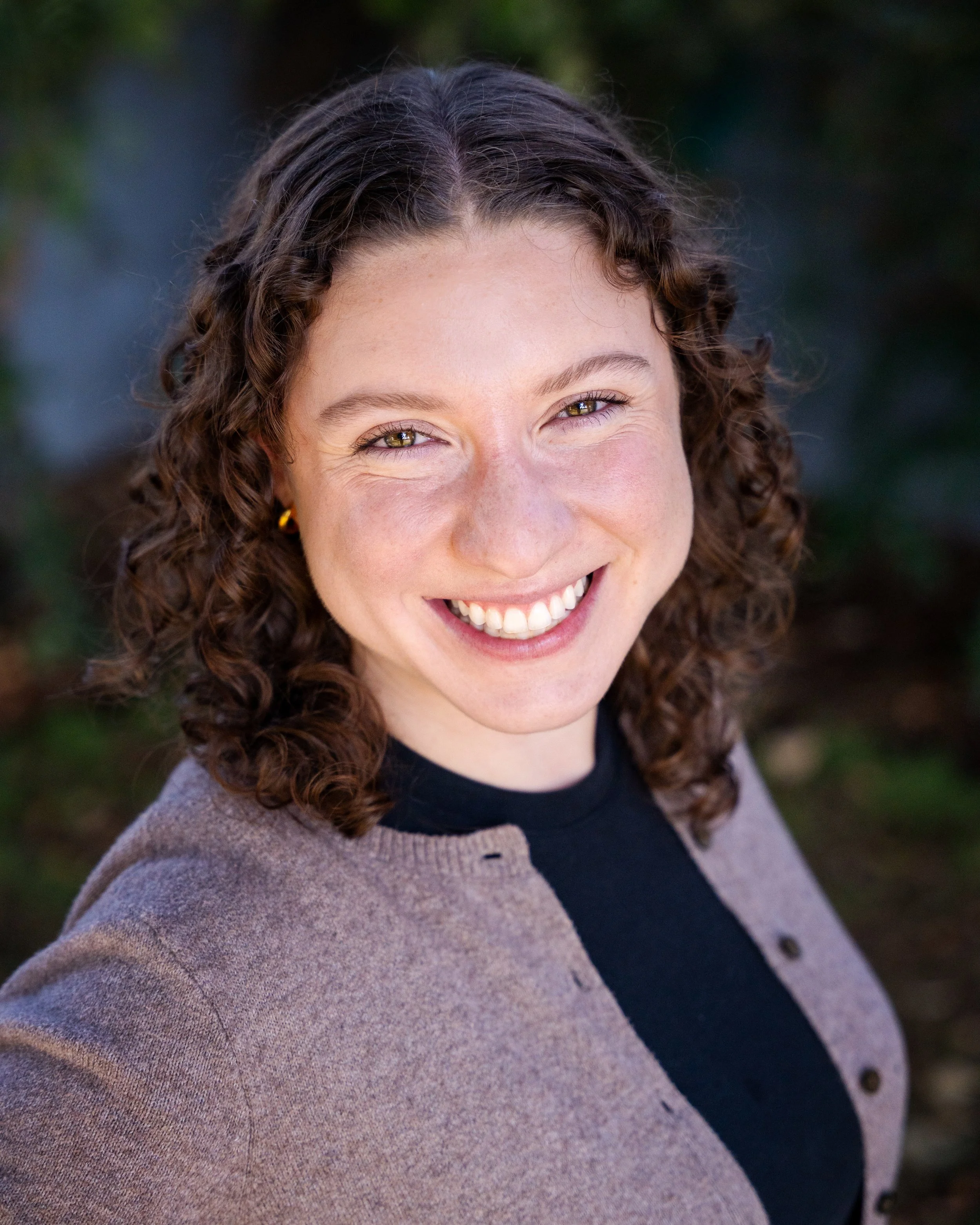 Close-up photo of a smiling woman with dark brown, wavy hair, wearing a white sweater, small hoop earrings, a nose piercing, and a delicate necklace with a small heart pendant, standing against a plain light gray background.