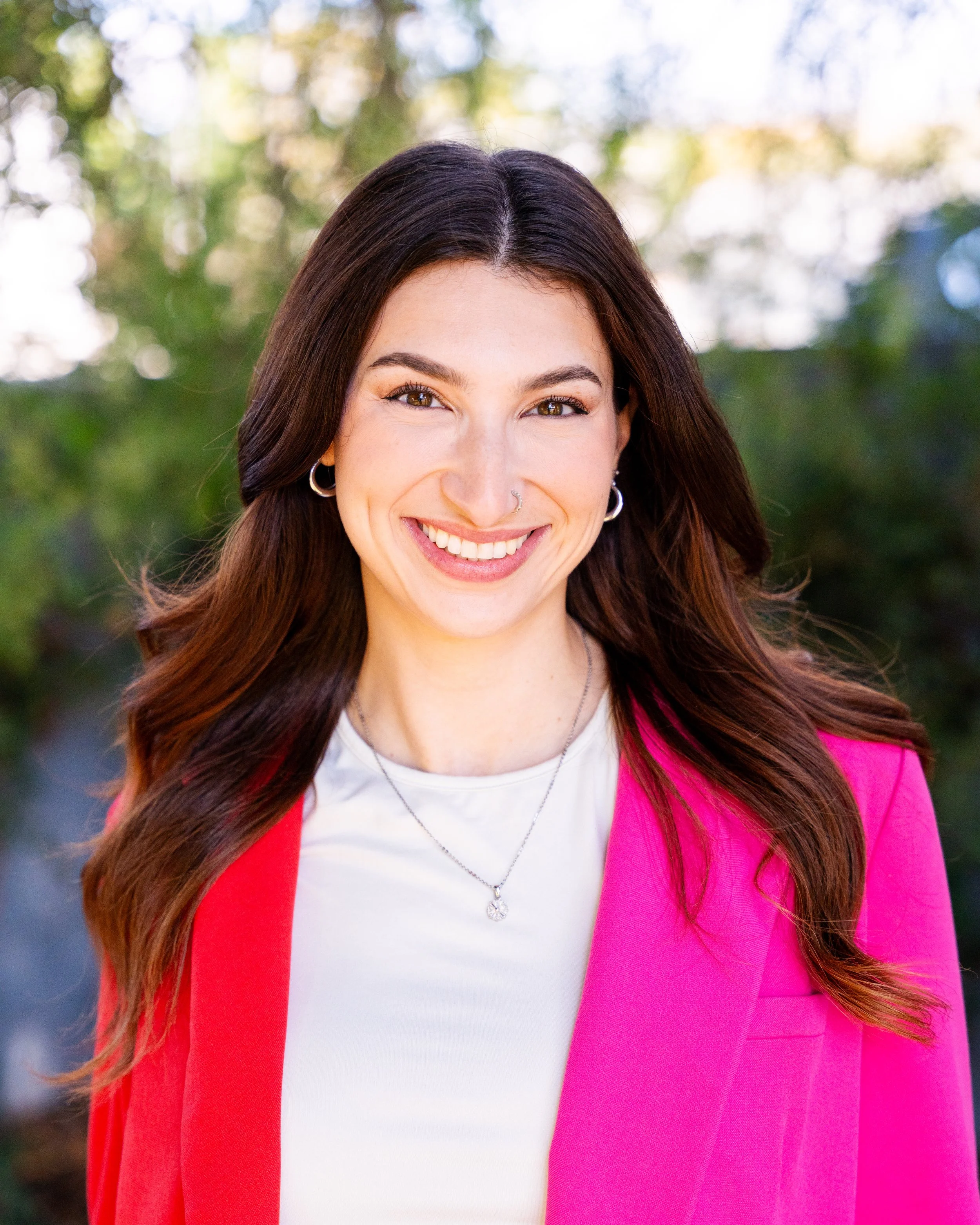 A woman with long, wavy, dark red hair, smiling and wearing a black blazer, white top, pearl necklace, and a nose piercing, standing against a wooden background.