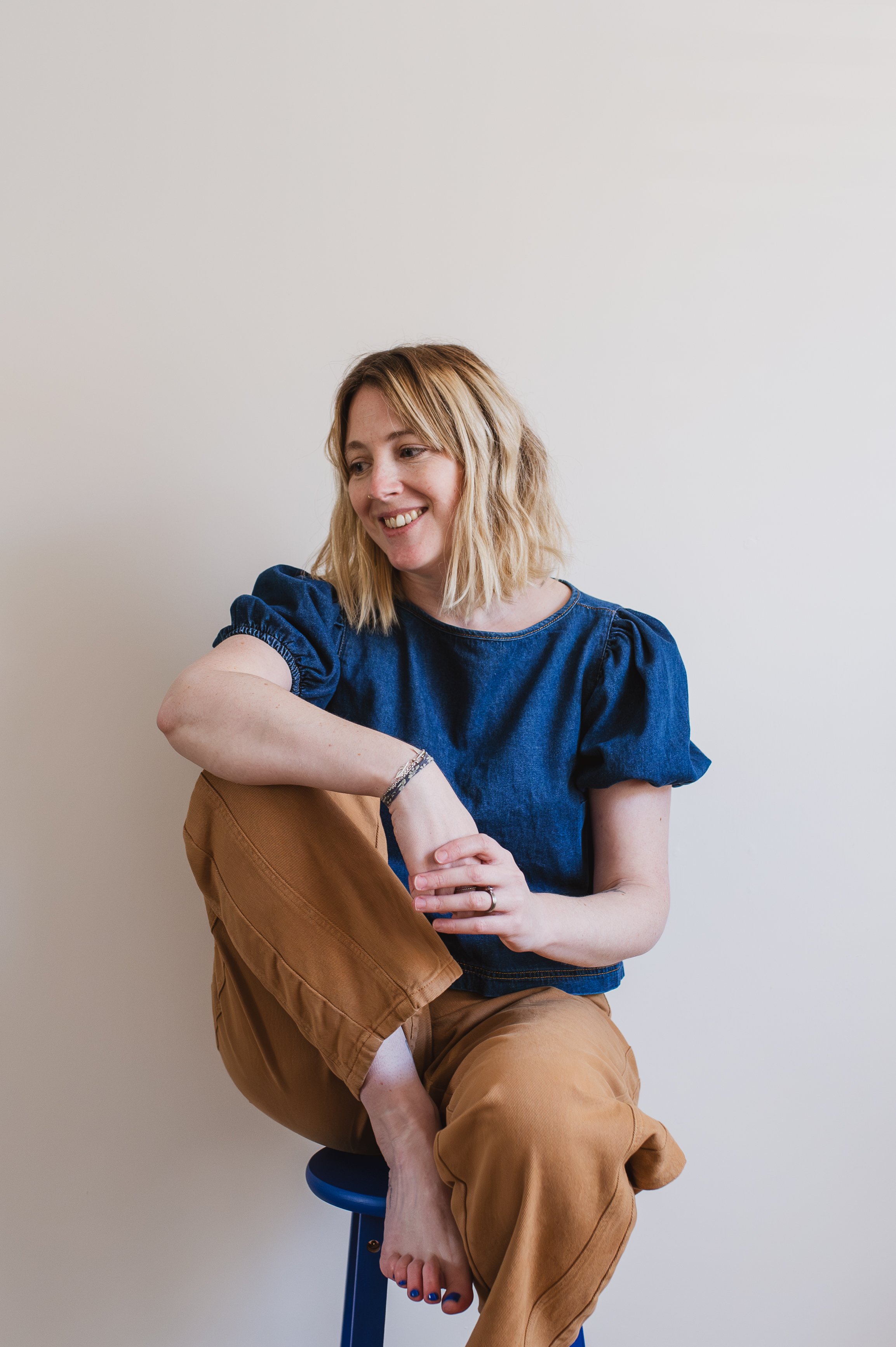  Lady with blonde hair sitting on a stall. She is wearing a blue top, tan trousers and no shoes or socks. 